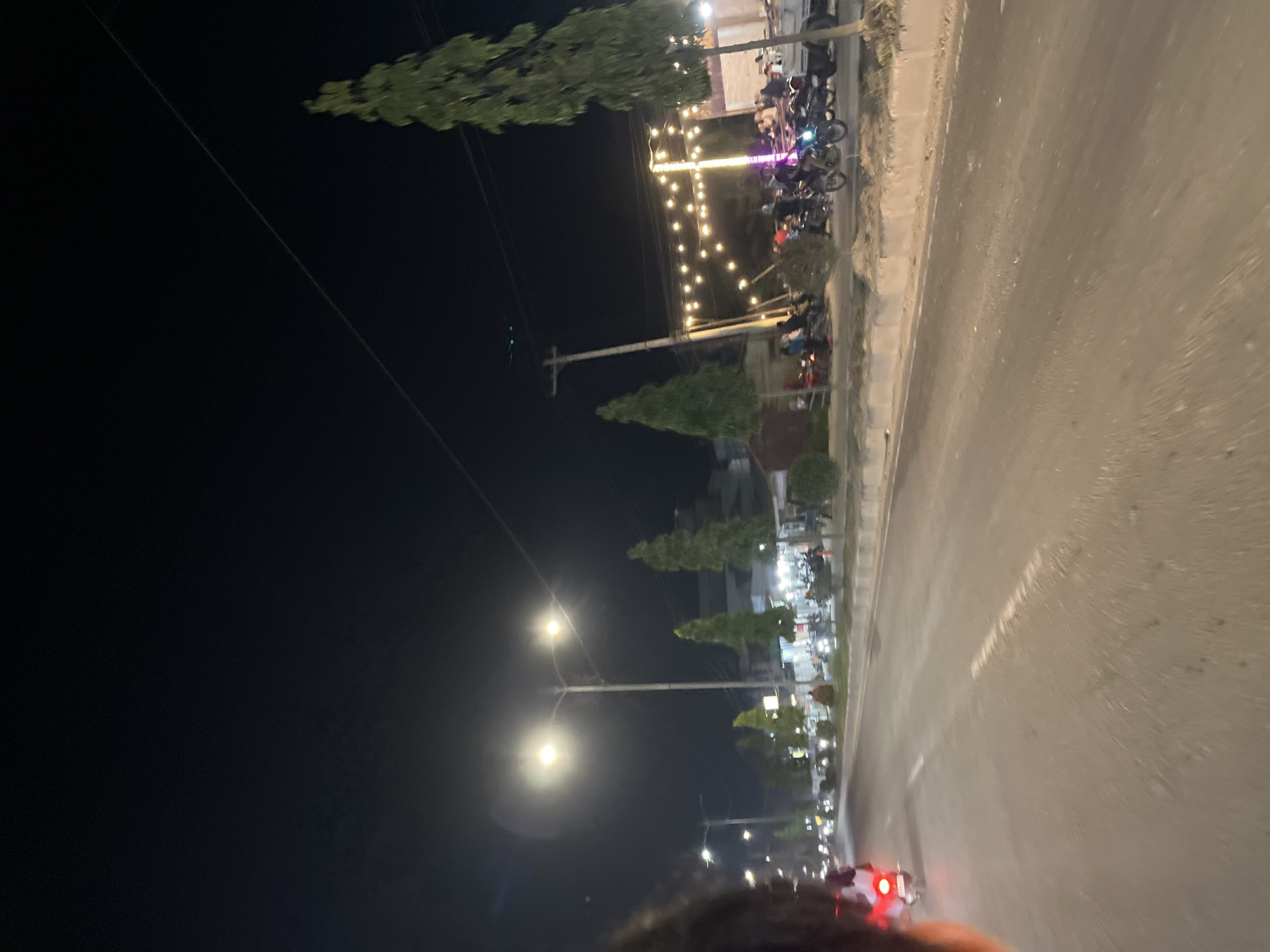 This media file depicts a nighttime scene on a street in Bundar, Indonesia. The perspective is from someone riding a motorcycle, as indicated by the red taillight visible in the foreground. The street is paved and appears wet, suggesting recent rain or ongoing damp conditions.
Along the side of the road, there are several parked motorcycles, indicating a gathering or a place of interest. A line of decorative string lights illuminates the scene, creating a festive and inviting atmosphere. Several trees, some conical in shape and others more mature and leafy, are planted along the roadside, adding greenery to the urban landscape. Streetlights are also present, casting bright pools of light.
In the background, buildings are visible, suggesting a commercial or residential area. People are present, some on motorcycles and others near the buildings, likely socializing or engaging in activities related to the establishments. The overall mood seems lively and social, despite the darkness of the night. The presence of motorcycles and the roadside setting are common in Indonesia. The time of day is clearly night, indicated by the darkness and artificial lighting.