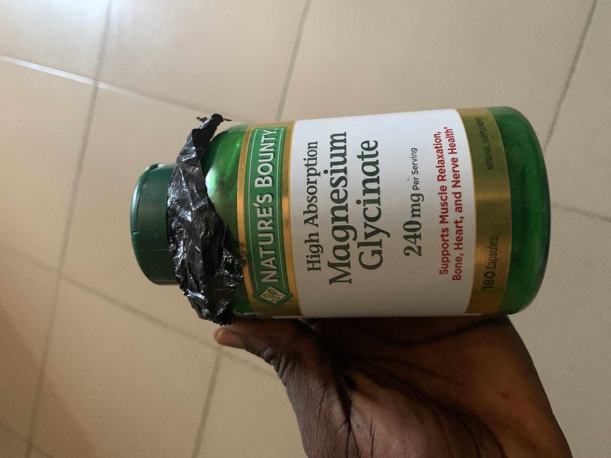 A person's hand with dark skin holds a green and white supplement bottle horizontally in the frame. The bottle is from the brand "NATURE'S BOUNTY" and contains "High Absorption Magnesium Glycinate." The label specifies "240 mg Per Serving" and states that it "Supports Muscle Relaxation, Bone, Heart, and Nerve Health*." It also indicates "180 Capsules" and that it is a "MINERAL SUPPLEMENT." The neck and cap area of the bottle are partially covered with a piece of crumpled black plastic or tape. The background consists of a light-colored, possibly tiled, floor with visible grout lines and a plain, light-colored wall, suggesting an indoor setting. The lighting is even, typical of an interior environment during the day.