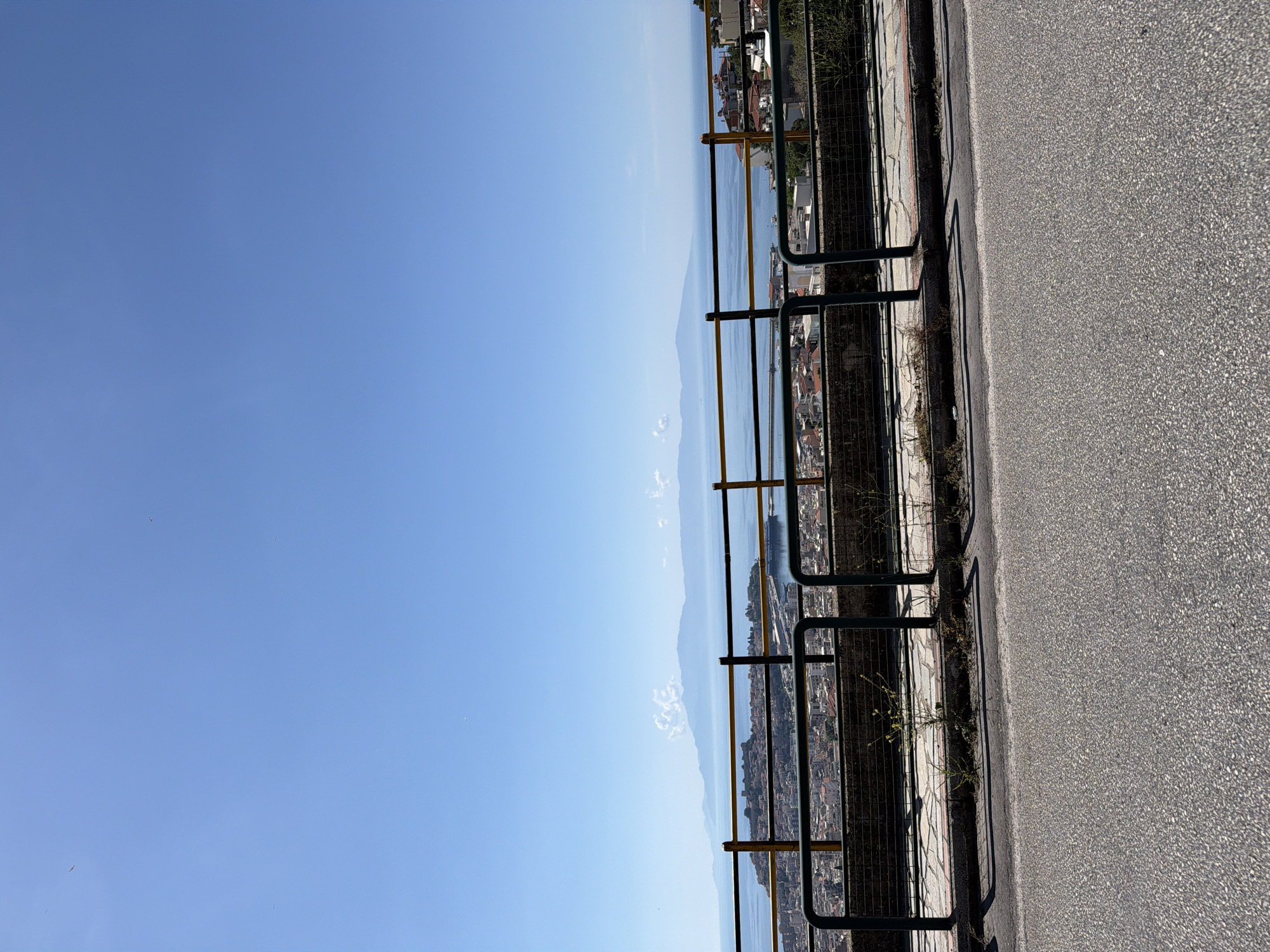 The image shows a view from a high vantage point in Kavala, Greece. A black and yellow metal railing runs along the edge of a paved road. Beyond the railing, there is a cityscape overlooking the Aegean Sea. Mountains are visible in the distance across the water. The sky is clear blue.