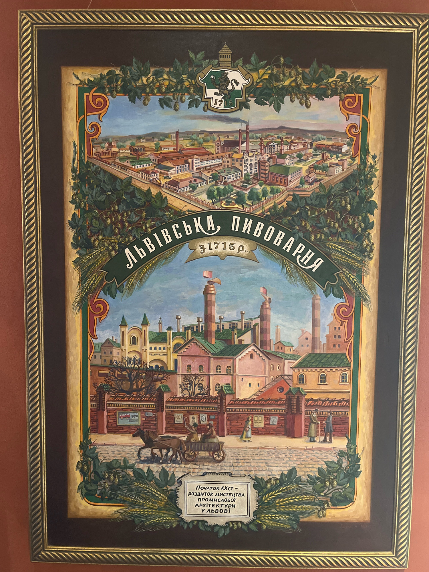 The image is a painting depicting an aerial view of a city with a brewery. In the upper portion of the painting, a panoramic view of a city with numerous buildings, chimneys, and industrial structures is shown. A banner with Cyrillic text and the year "1715" is prominently displayed across the middle of the painting. Below this banner, a closer view of the brewery complex is depicted. This section shows several brick buildings with green roofs, tall smokestacks, and architectural details. In the foreground of this lower section, a horse-drawn cart with figures is on a cobblestone street. Two figures stand on the sidewalk near the buildings. At the very bottom, a rectangular plaque contains Cyrillic text. The painting is framed by a decorative border featuring intertwined leaves and hops, as well as ornate gold detailing. The overall style is illustrative and historical. Based on the text visible on the banner, "ЛЬВІВСЬКА ПИВОВАРНЯ 1715р.," the painting likely represents the Lviv Brewery in Lviv, Ukraine, and its founding year. The plaque at the bottom also contains text that likely provides further historical context about the brewery or its architectural significance.