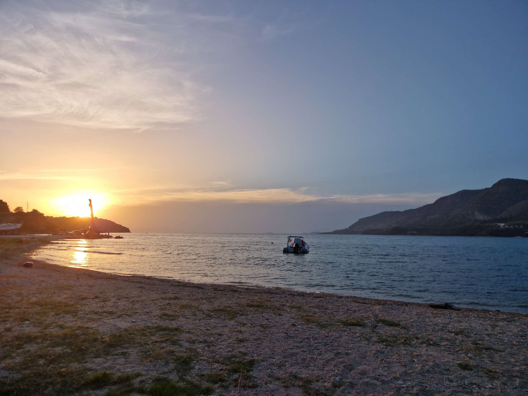 A wide-angle photograph captures a serene sunset scene at the beach in Plataria, Greece. The sky is a gradient of soft orange and pink near the horizon, transitioning to a clear blue higher up, with wispy clouds scattered throughout. The sun, a bright disc low on the horizon, casts a shimmering reflection across the calm sea. In the foreground, a pebble beach is visible with patches of dry grass. A small, white boat with a blue stripe and two people on board floats gently on the water. To the left, a construction crane and some structures are visible on the distant shoreline. The background is dominated by the dark silhouette of a mountain range. The overall impression is one of tranquility and natural beauty.