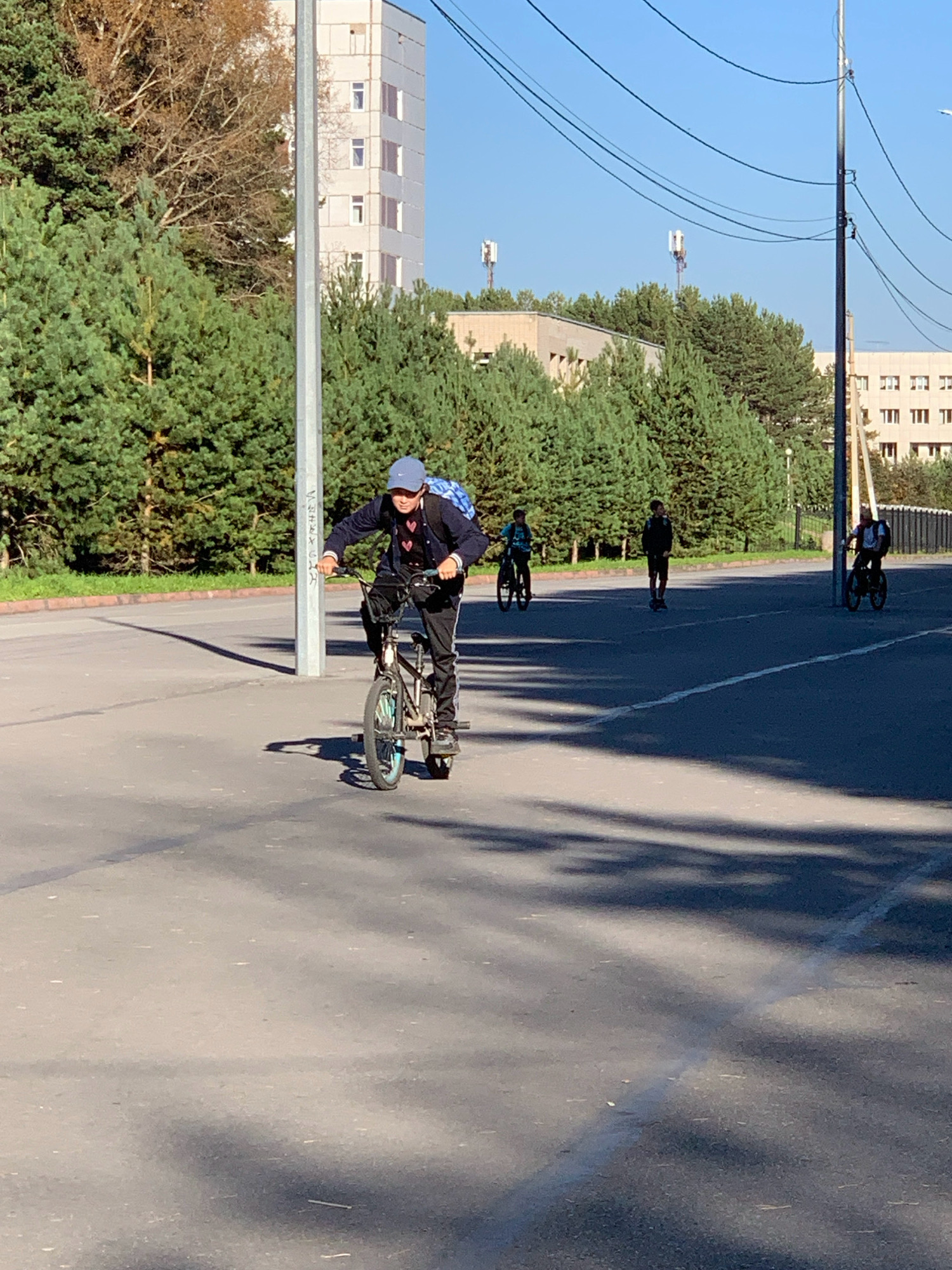 The image captures a young male on a BMX bike, wearing a blue baseball cap and a backpack, riding on an asphalt road. He is positioned in the foreground, facing slightly to his left, with his right hand on the handlebars. The bicycle has a blue rim on the front wheel and pegs on the rear axle. In the middle ground, two other individuals are visible; one on a bicycle and another standing. Further in the background, there are trees, buildings, power lines, and two cell towers. The scene suggests an outdoor setting in a city, likely a public space like a park or street. The time of day appears to be late afternoon or early evening, indicated by the long shadows cast on the road. The location is identified as Prokopyevsk, Russia.