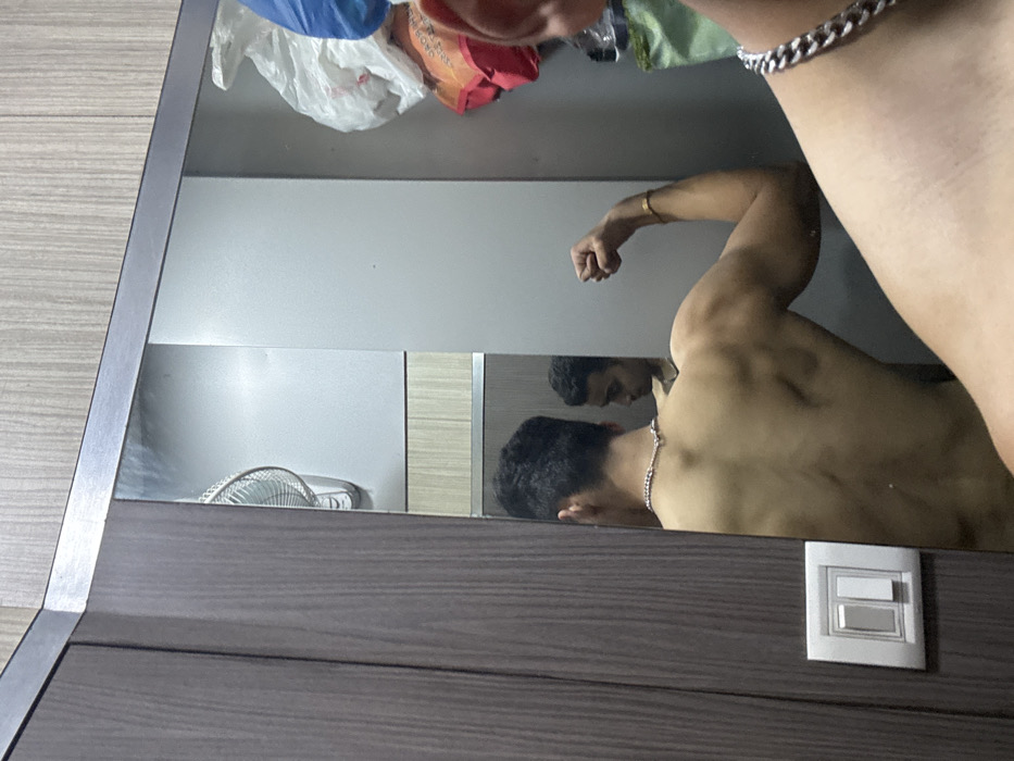 The scene is an indoor setting within the city of Kolkata, India, featuring a male subject captured in a mirror selfie. The male, bare-chested, is visible from the upper chest to mid-torso, wearing a silver-colored chain necklace. His back is turned towards the viewer, and his reflection in the mirror shows him from the back, posing with his left arm flexed, displaying his bicep. A gold-colored bracelet is present on his left wrist. His face is visible in profile in the reflection, looking towards his left arm.
The environment includes a large mirror, partially obscured on the left by a dark brown wood-textured wardrobe or cabinet. A white light switch panel with two rectangular switches is mounted on the wall or cabinet to the left of the mirror. Reflected in the mirror, behind the subject, are light-colored walls, with a section of wood-grain textured paneling below. A white wall-mounted oscillating fan is partially visible in the upper-left of the mirror's reflection. In the upper-right portion of the reflection, various items are visible, including a white plastic bag, a blue fabric item, and an orange/red fabric item, suggesting a lived-in private space.