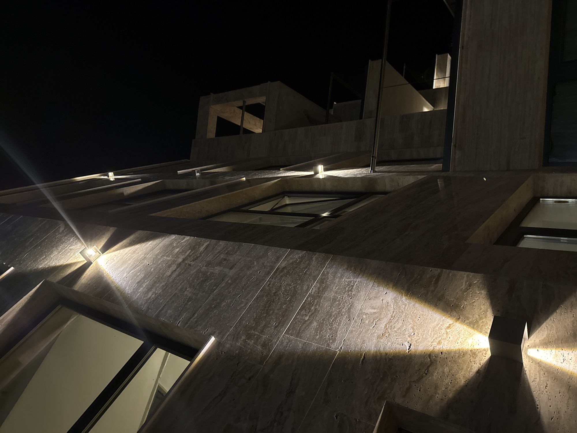 The image shows the exterior of a modern building at night in Tyulenovo, Bulgaria. The building features a facade clad in light-colored, horizontally striated stone or tile. Several exterior lights are mounted on the wall, casting dramatic upward and downward beams of warm light. These lights illuminate portions of the stone, creating contrasting areas of light and shadow. In the background, the dark night sky is visible, with no stars or moon apparent. The architecture includes geometric shapes and what appears to be a balcony or open-air section with a glass railing. One section of the building features an angular, abstract architectural element that is also illuminated by the lighting. The overall impression is one of modern, minimalist design emphasized by dramatic nighttime illumination. There are no people or animals visible in the image.