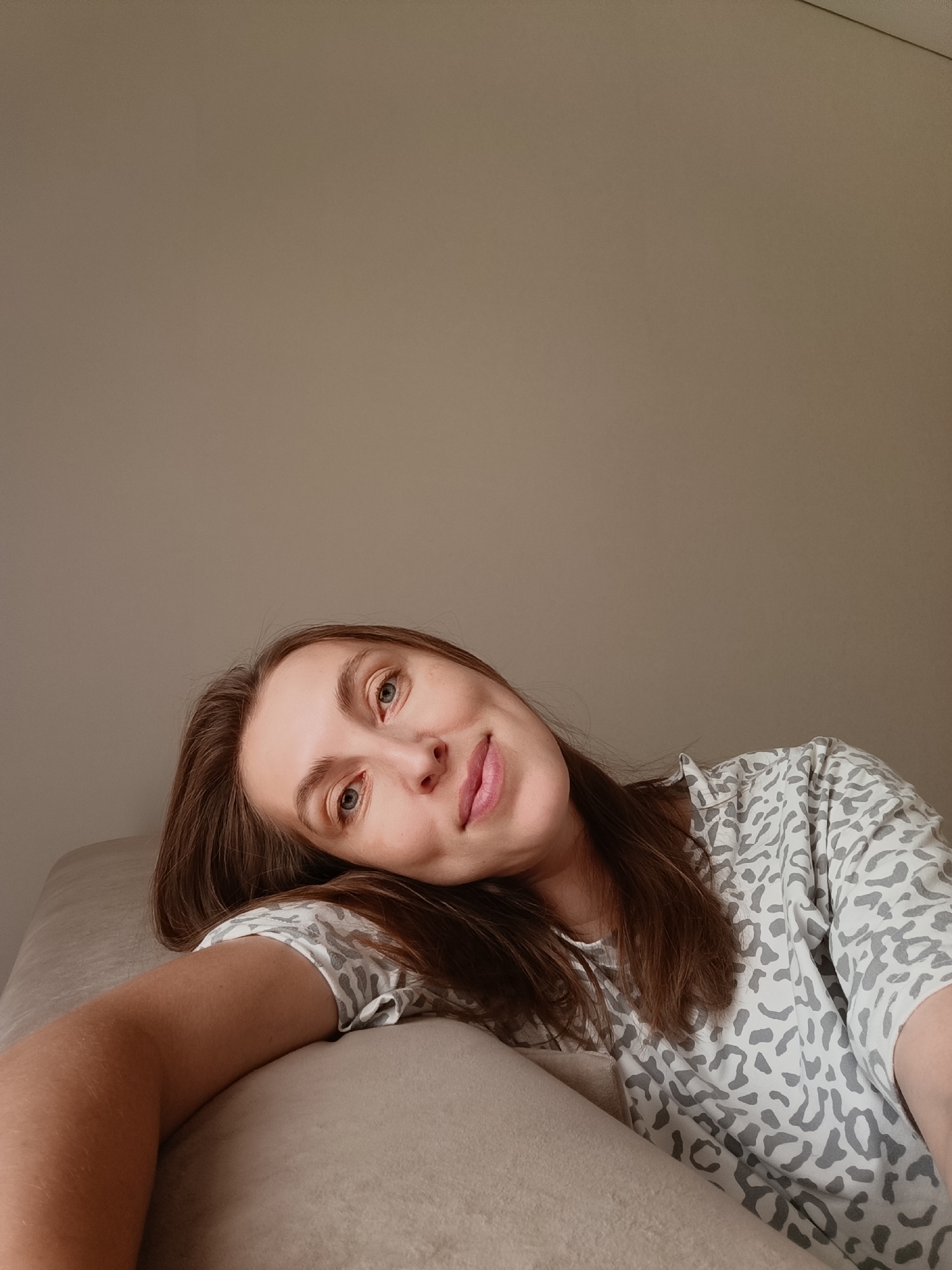 A young woman with long, brown hair is captured in a selfie, resting her head on a beige surface. She is wearing a gray and white patterned t-shirt. Her head is tilted, and her gaze is directed towards the camera with a playful expression, her tongue slightly sticking out. The background is a plain, light brown wall, suggesting an indoor setting. The lighting is soft, implying it could be daytime with diffused sunlight or indoor lighting. The overall mood of the image is casual and relaxed. There is no visible text in the image. The location context is Chaikovsky, Russia.
