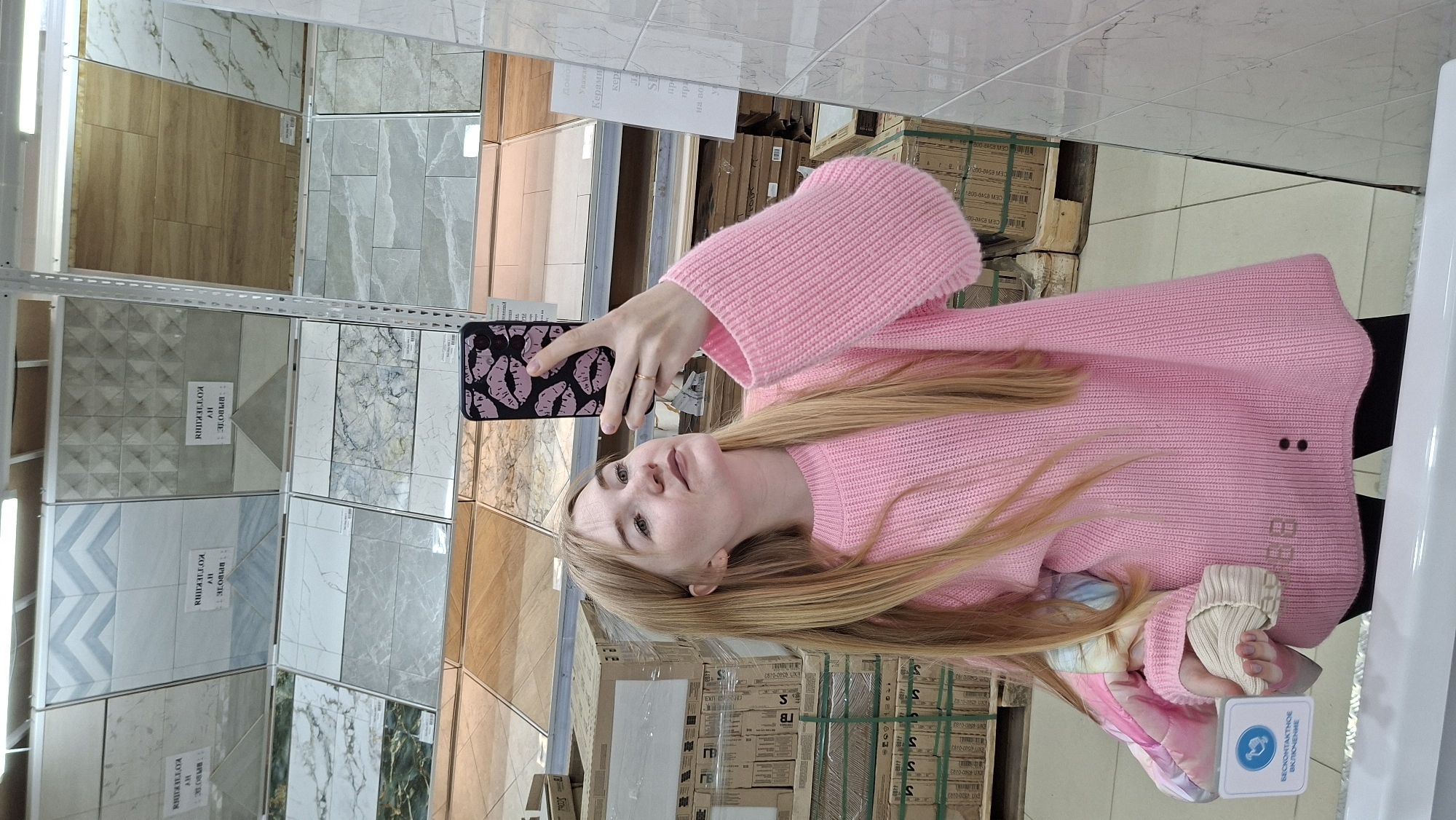 A young woman with long blonde hair is taking a selfie in a retail store, seemingly a tile shop. She is wearing a pink sweater and holding a smartphone with a black case decorated with pink lips. Behind her are shelves displaying various tile samples, some with Russian labels (e.g., "KOTIERIIH"), and boxes of tiles are visible in the background. The location is potentially in Chaikovsky, Russia.