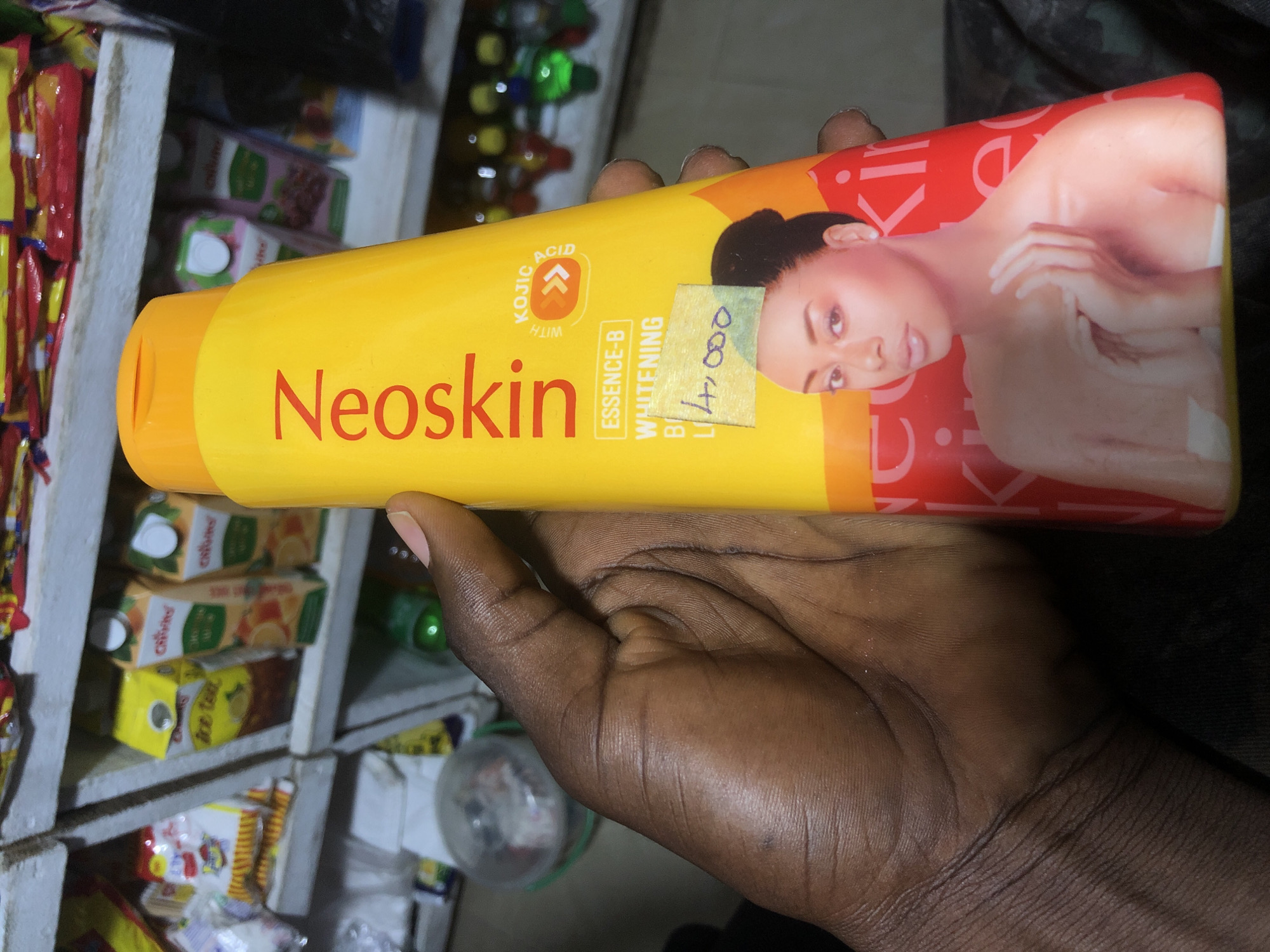 A person's hand holds a yellow bottle of Neoskin Essence-B Whitening Body Lotion. The bottle has a picture of a woman on it and text that reads "WITH KOJIC ACID". A handwritten price tag of "14,000" is affixed to the bottle. The background appears to be a store shelf with various products, including juice boxes and bags of snacks. The scene is likely a market or convenience store in Ado, Nigeria, given the brand and currency.