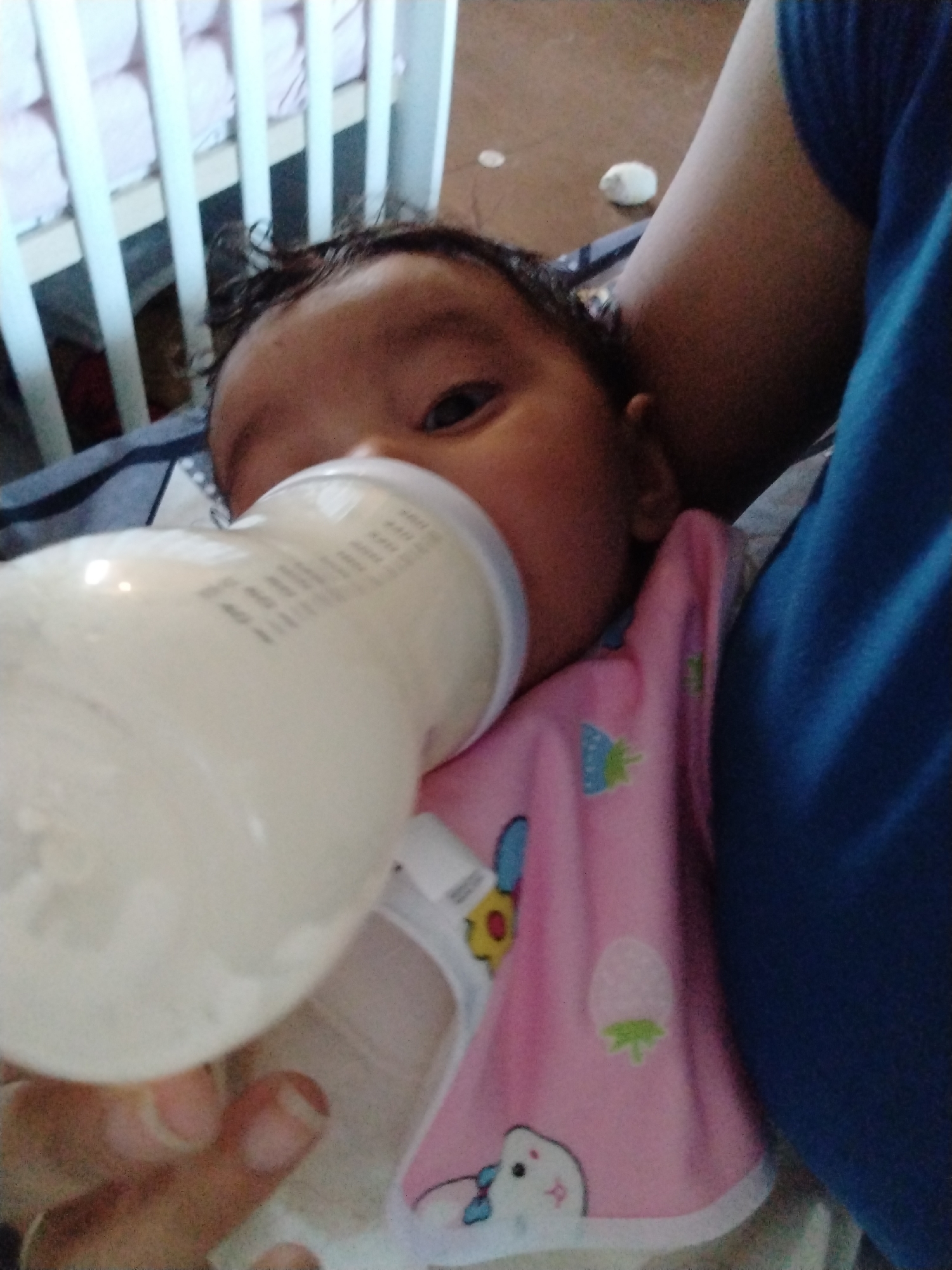 A baby is being bottle-fed in an indoor location, near a crib. The baby is wearing a pink bib with cartoon characters. A person in a blue shirt is holding the baby. The location is unspecified.