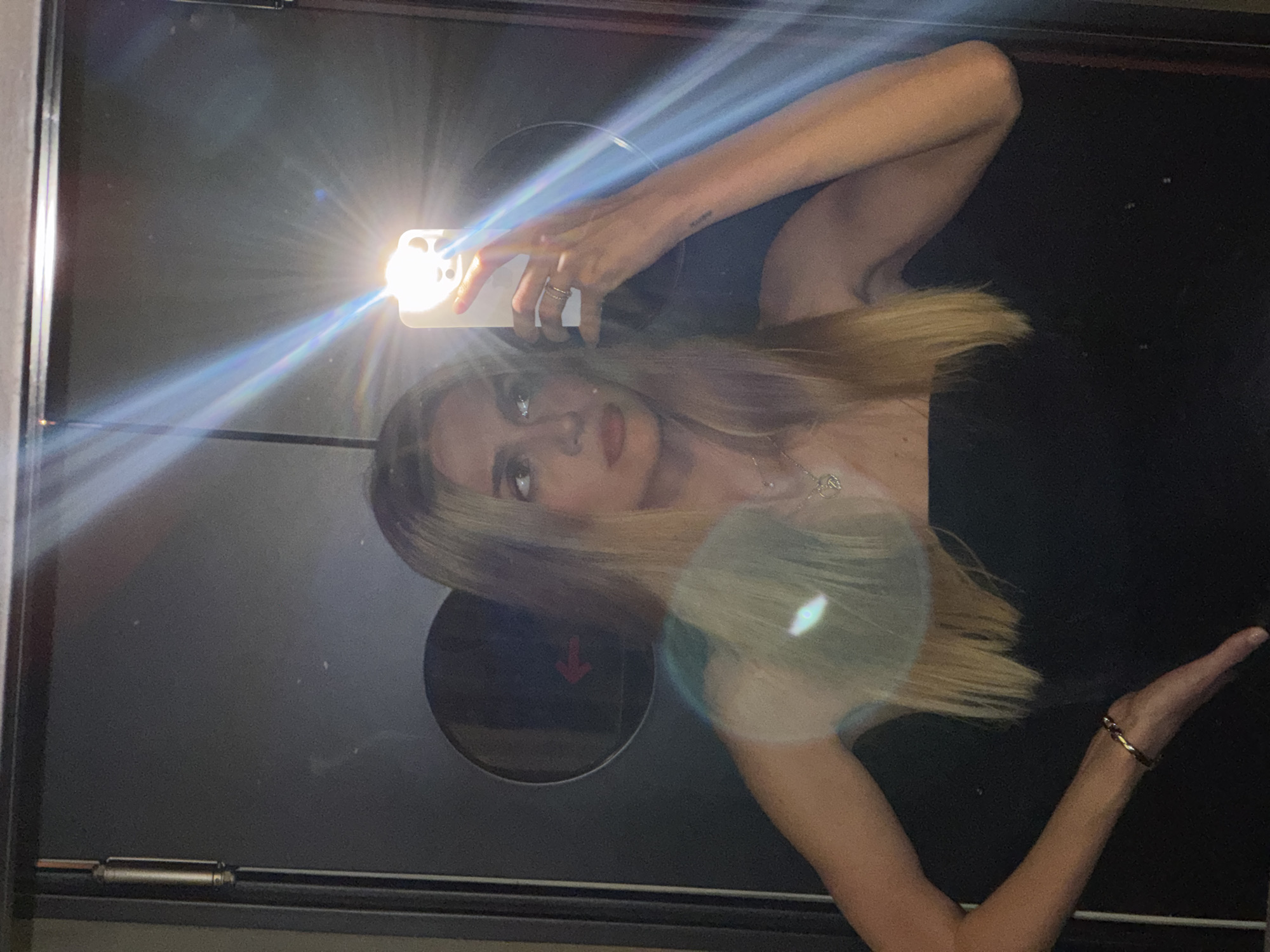 A young woman with long blonde hair is taking a selfie in a mirrored elevator. She is wearing a black top and jewelry, including a necklace and bracelet. The elevator door has a circular indentation with a red arrow inside. The location is likely in Tbilisi, Georgia.