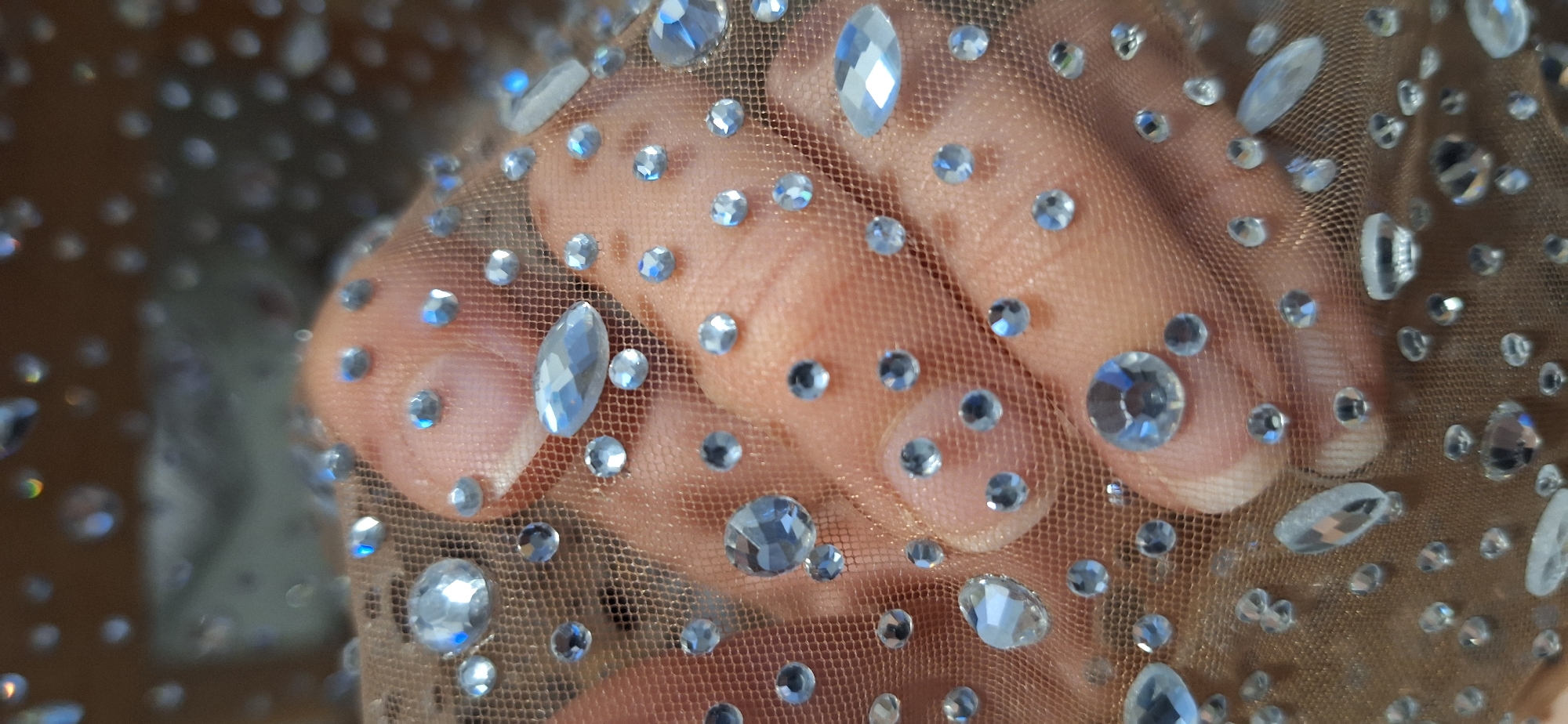 A close-up shot shows a hand covered with a layer of sheer fabric. The fabric is adorned with numerous small, clear rhinestones or crystals that catch the light. The rhinestones vary in size and shape, with some being round and others elongated. The fabric appears to be stretched or gathered over the hand, creating texture. The hand's skin is visible through the fabric, and the overall effect is glittering. There is no visible indication of the location being Blagoevgrad, Bulgaria.