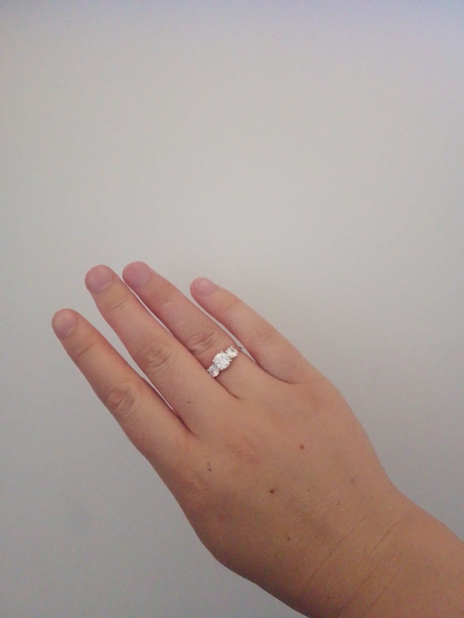 A hand, adorned with a three-stone ring, rests against a plain, light-colored background. The ring features a central, larger stone flanked by two smaller stones, all appearing to be clear and faceted. The hand is positioned with the palm facing upwards and slightly angled away from the viewer, revealing the back of the fingers. The fingers are relaxed and slightly curved. The background is a uniform, neutral tone, lacking any discernible features or context that would place the image in a specific city or location. No elements in the image suggest a connection to Arbagish, Uzbekistan.