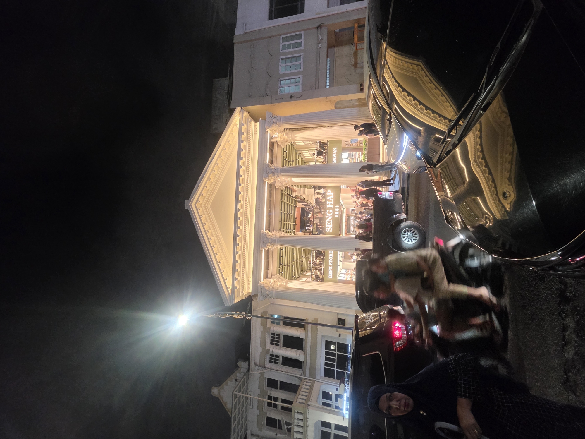 The image captures a nighttime scene in Medan, Indonesia, focusing on a brightly lit department store with a classical facade. The store, identified by signs reading "SENG HAP 1881" and "SAWAN," features grand columns and illuminated architectural details, suggesting a grand opening or a busy shopping evening.
Several people are visible, both inside and outside the store, with some browsing merchandise and others interacting on the sidewalk. A black car is parked in the foreground, partially obscuring the view of the building and reflecting the store's lights. Another vehicle, possibly a motorbike, is also present, with a blurry figure on it. The overall atmosphere appears lively, with the warm lighting of the store contrasting with the dark night sky. A street lamp shines brightly in the upper left quadrant of the image, adding to the nighttime illumination. The architecture of the building, with its prominent columns and pediment, gives it a sense of historical significance. The presence of people and vehicles suggests an active urban environment.