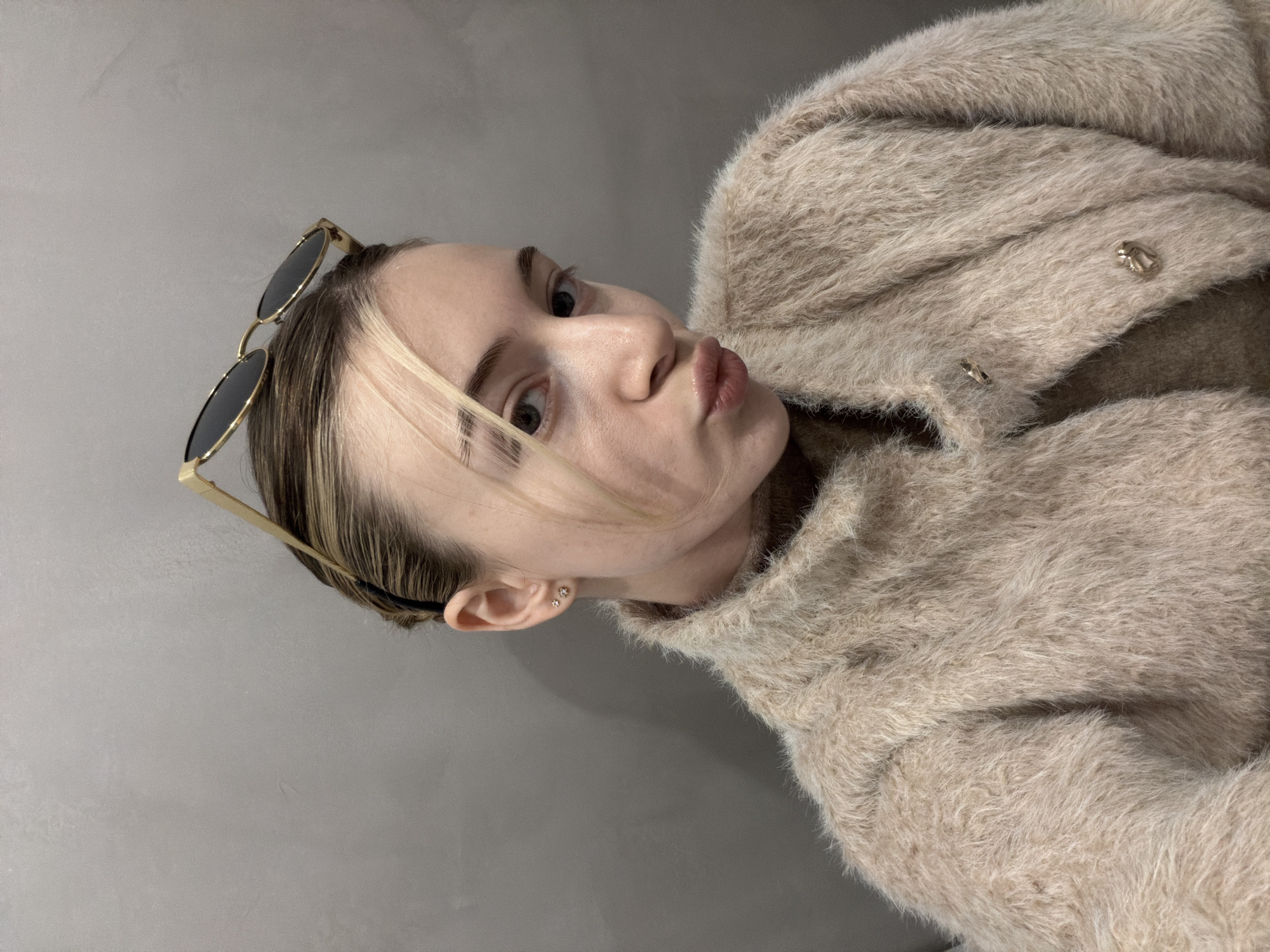 A young woman is posing for a selfie indoors against a plain gray wall. She has fair skin, light brown hair with a blonde streak, and is wearing gold-rimmed sunglasses pushed up on her head. Her lips are puckered slightly. She is wearing a fluffy, light brown sweater or jacket with button closures. The location is unknown.