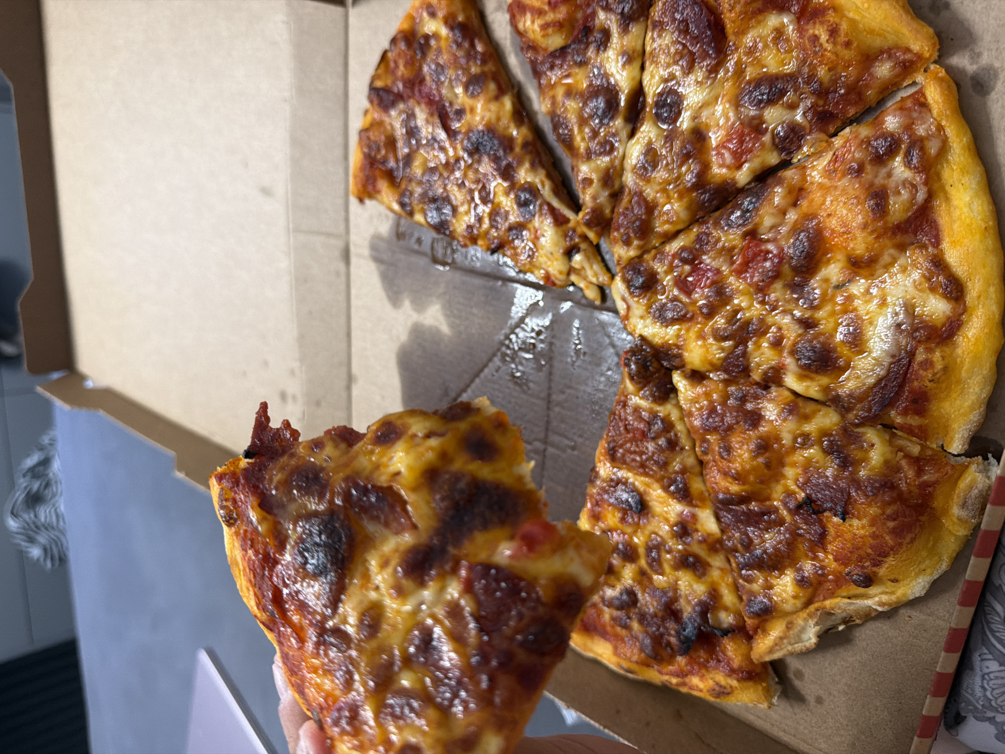 A hand holds up a slice of pepperoni pizza, with more slices of pizza inside a cardboard pizza box. The pizza has a golden crust, melted cheese, and red sauce. The background is blurred and shows a table and a gray surface. The scene does not provide any information about the city of Ruse, Bulgaria.