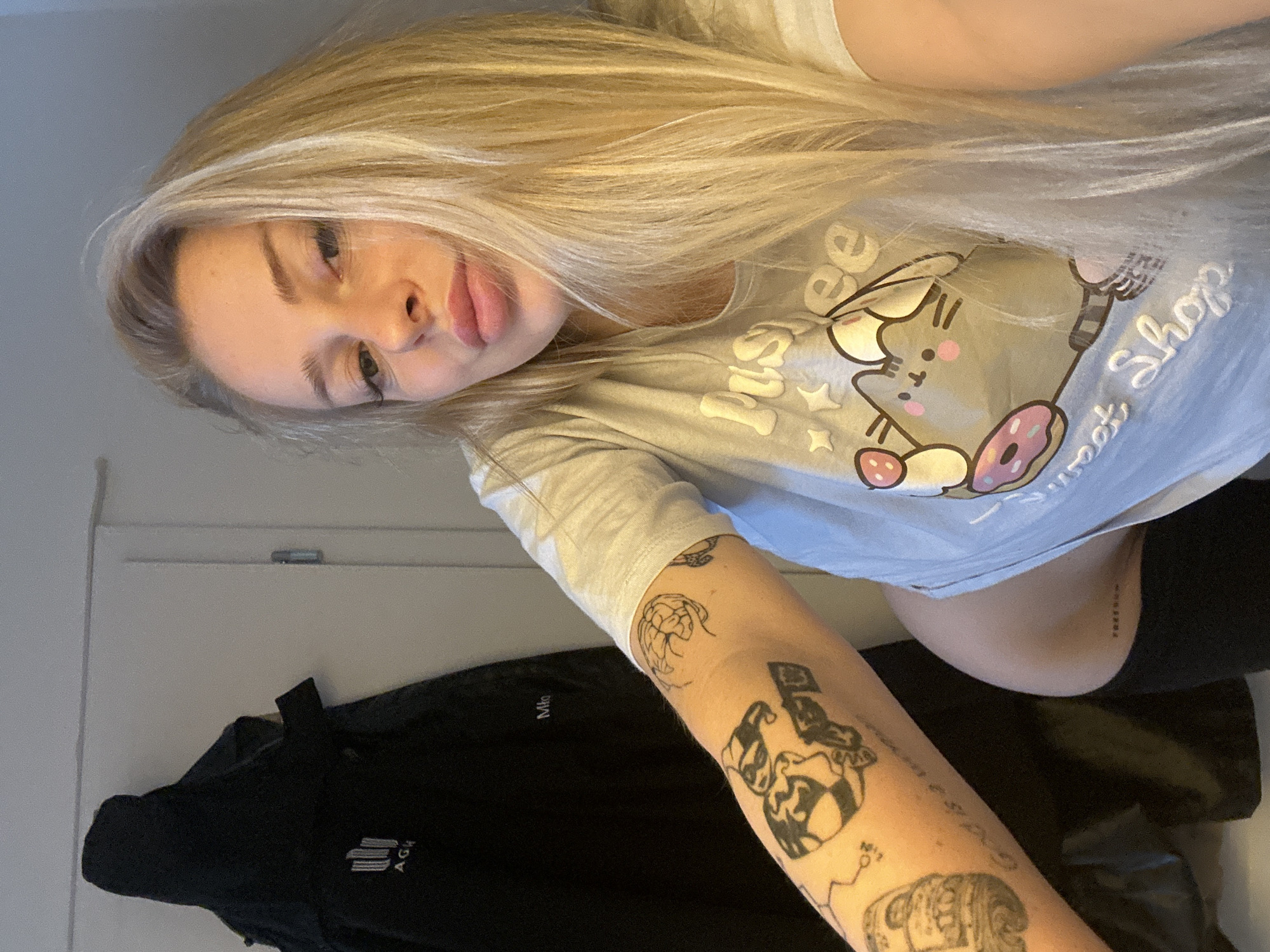A blonde woman with tattoos on her arm poses for a selfie in an indoor setting. She is wearing a light-colored t-shirt with a cartoon cat graphic and text that reads "Pusheen Sweet Shop". Her left arm, which has several tattoos, is extended towards the camera. In the background, a black jacket is hanging on a wall. The image appears to be taken in an urban residential environment, possibly an apartment. There is no information to suggest the location is Krakow, Poland.
