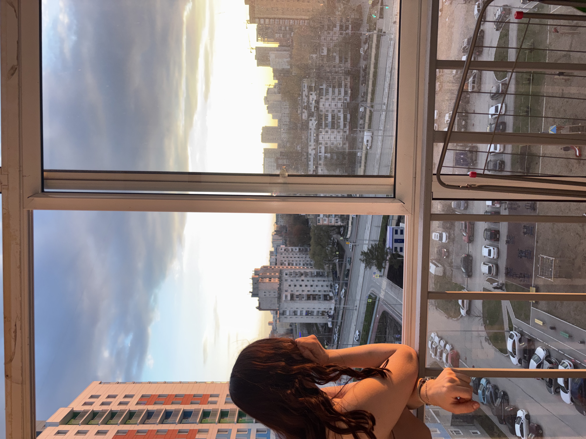 A person with long, dark, wavy hair is observed from behind, looking out a large, multi-pane window, likely from a high-rise building or apartment in Perm, Russia. The individual's shoulders and back are partially visible, and a thin bracelet adorns their right wrist.
The expansive window offers a broad view of an urban landscape, characterized by numerous multi-story buildings. Some structures appear older, while others show signs of ongoing construction. A street is visible below, bustling with parked and moving cars. A metallic clothes drying rack on a balcony partially obstructs the view of the street on the right side of the frame.
The scene is bathed in the warm, golden light typical of late afternoon or early evening, suggesting a sunset or sunrise. The sky features a mix of bright areas and clouds. A small bottle rests on the windowsill between the central window panes.
Several architectural elements stand out, including multi-colored window accents (green/blue and orange/red) on a large building in the lower left. Road markings and pedestrian crossings are discernible on the street. A sign with unidentifiable Cyrillic text is visible on the street level, near parked vehicles, specifically a white sign with dark text. The overall view captures a snapshot of daily life in a bustling urban environment.