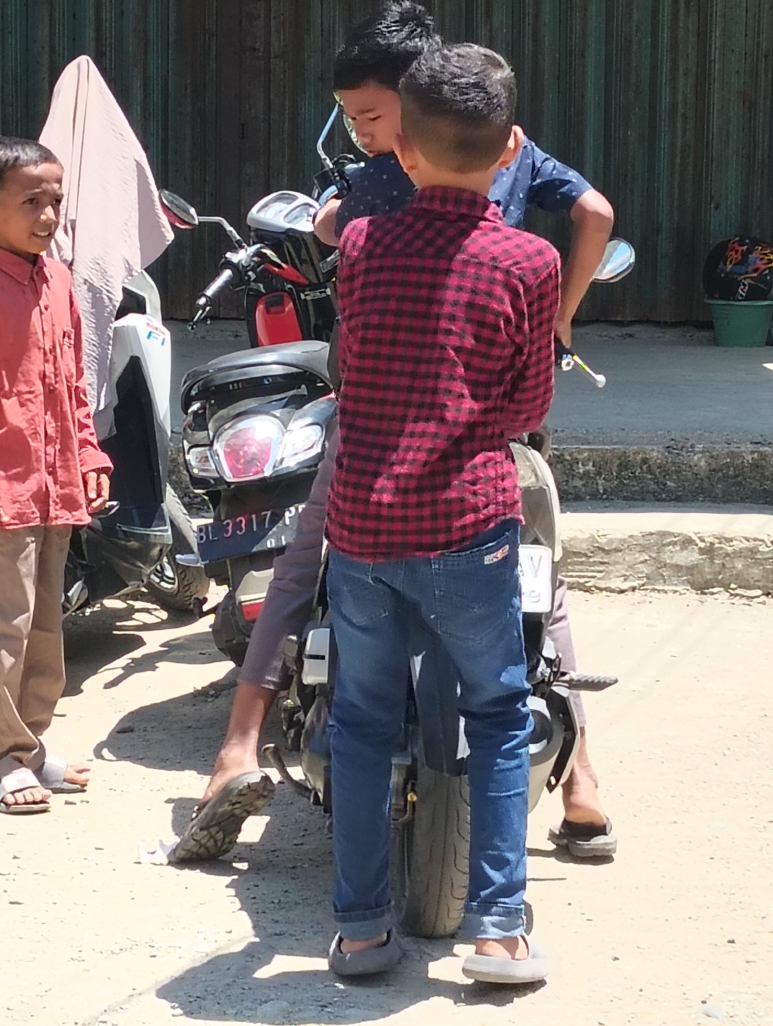 In Teungoh, Indonesia, three young boys stand near motorcycles on a sunlit dirt road. One boy in a red and black checkered shirt and blue jeans is positioned centrally, facing away from the camera, with his hands on a motorcycle's handlebars. To his left, another boy wearing a blue shirt with a white pattern is partially visible, also near the motorcycles. On the far left, a boy in a reddish-orange collared shirt and light-colored pants stands looking to the left. A motorcycle's rear is prominent in the foreground with license plate "BL 3317 PR". In the background, a green corrugated metal wall forms the backdrop, with a green bucket containing a helmet visible on the right. The environment is dusty, with shadows indicating a bright day.