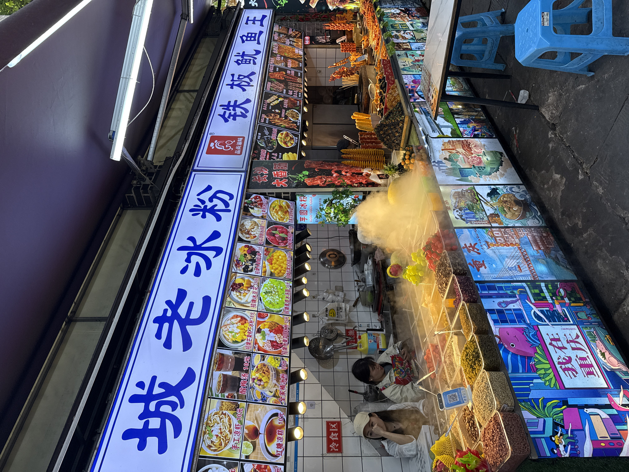 Here is a detailed description of the image:
**Scene:** A bustling street food stall, likely at night, possibly located in the Nan'an District, Chongqing, China. The stall is selling various food items, including snacks, sweets, and drinks.
**People:** Two women are seen inside the food stall. Both appear to be wearing white coats or shirts, likely working there.
**Subjects:** Food items are the main subject. Signs show menu items like "Bingfen" (ice jelly), fruits, snacks, and cooked meats. There's also a sign for "Tieban Youyu Wang" (Grilled Squid King).
**Objects:**
* Food preparation equipment: Pots, pans, and serving utensils are visible.
* Menu signs: Displaying images and prices of the food.
* Ingredients: Visible in containers, including beans, fruits, and toppings.
* Tables and stools: Provide seating for customers.
* Lighting: Fluorescent lights illuminate the stall.
**Actions:** The food is displayed, prepared, and likely being sold. The people are working.
**Interactions:** Interactions between customers and vendors are likely taking place.
**Environment:** The stall is brightly lit and filled with food. Steam rises from the food. The area is crowded, suggesting a popular location. The environment appears to be urban.