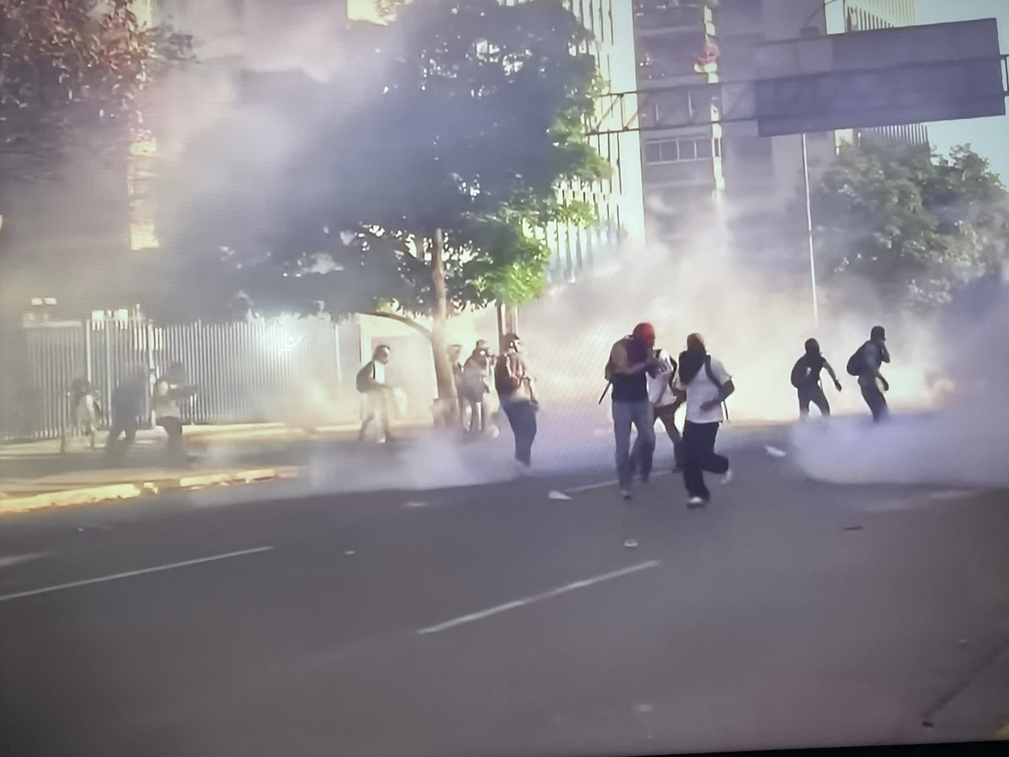 A street scene in an urban area with a group of people moving through a cloud of what appears to be smoke or tear gas. There are buildings in the background, and trees along the side of the road. The people are moving away from something in the background.