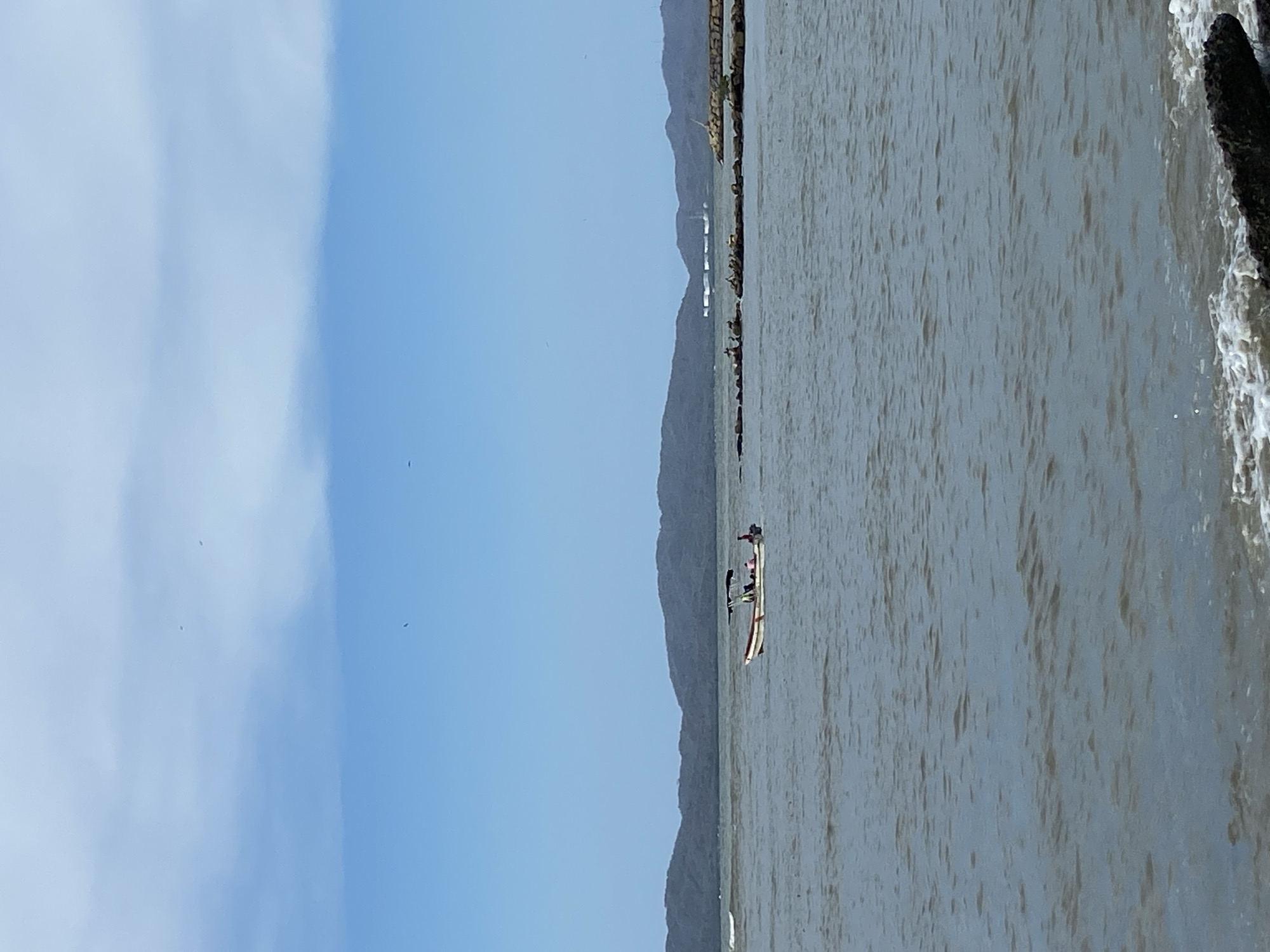 The image depicts a coastal scene, possibly in or near Higuerote, Venezuela, given the landscape and local boat. The right portion of the image shows a body of water with visible waves lapping against a dark, rocky shore. In the center of the frame, a narrow boat, likely a dugout canoe or similar small watercraft, is visible with what appear to be people aboard. In the background, there are a series of low mountains. The left portion of the image features a clear sky with some cloud cover. The image appears to have been rotated 90 degrees counterclockwise.