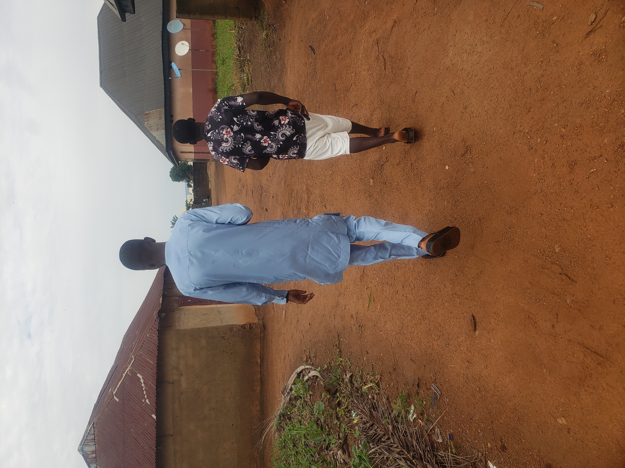 Two young Black males are walking away from the camera down a dirt path in what appears to be a rural area or the outskirts of a town. The male on the left is wearing a light blue traditional outfit, while the male on the right is wearing a black shirt with a floral pattern and white shorts. The environment consists of two buildings with corrugated metal roofs and what look like satellite dishes mounted on the side of one of the buildings. The ground is reddish-brown dirt, and there is some vegetation along the left side of the path. The sky is overcast.