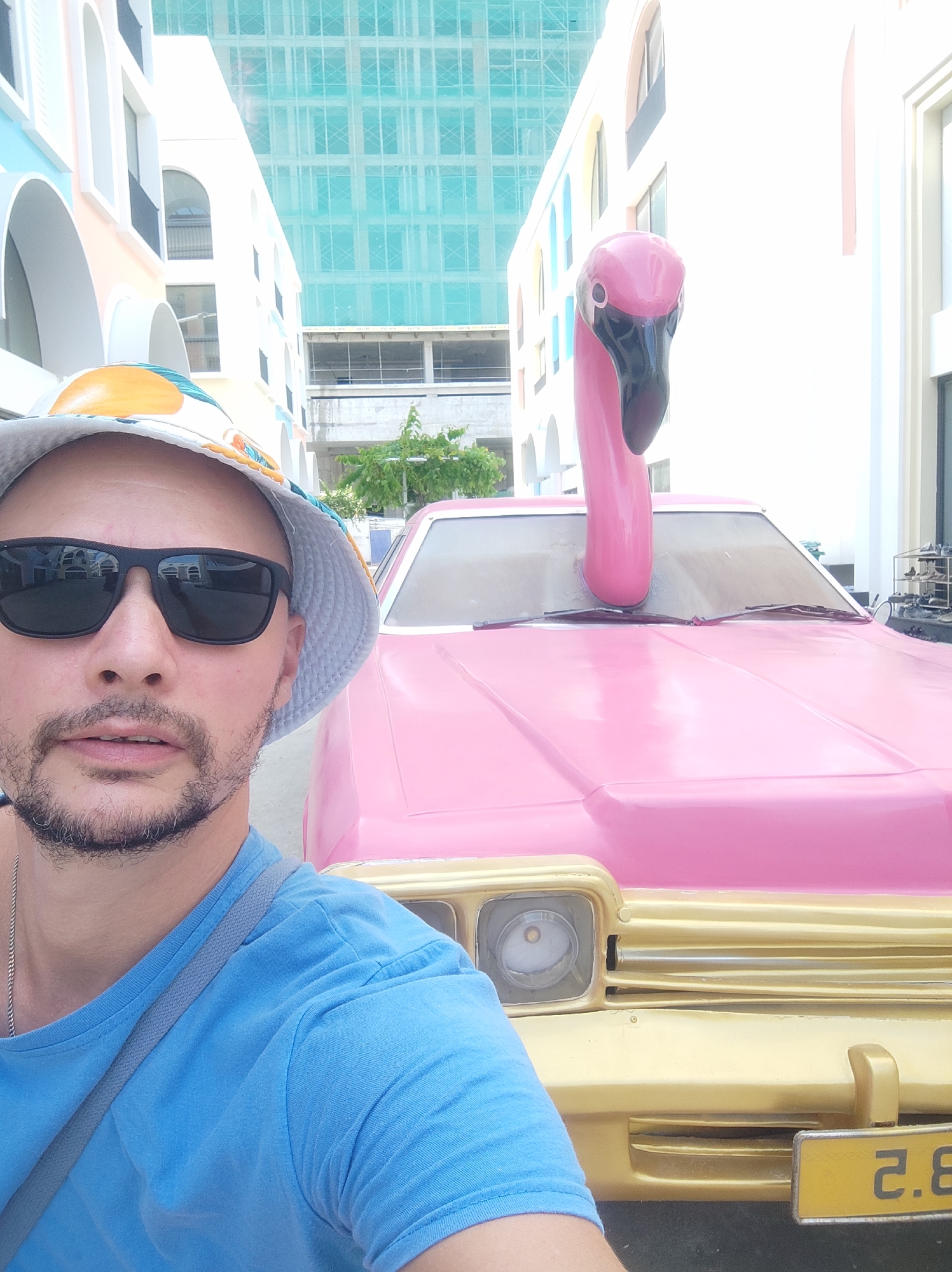 A man wearing a blue t-shirt, a bucket hat, and sunglasses is taking a selfie in front of a pink car with a flamingo statue protruding from its hood. The car is parked in an urban environment, with colorful buildings and a construction site visible in the background. The man is positioned in the foreground, with the car and its unusual adornment dominating the mid-ground and background of the image. The street and pavement are visible at the bottom of the frame. The scene appears to be a public space within the city of Bac Nha Trang Ward, Vietnam.