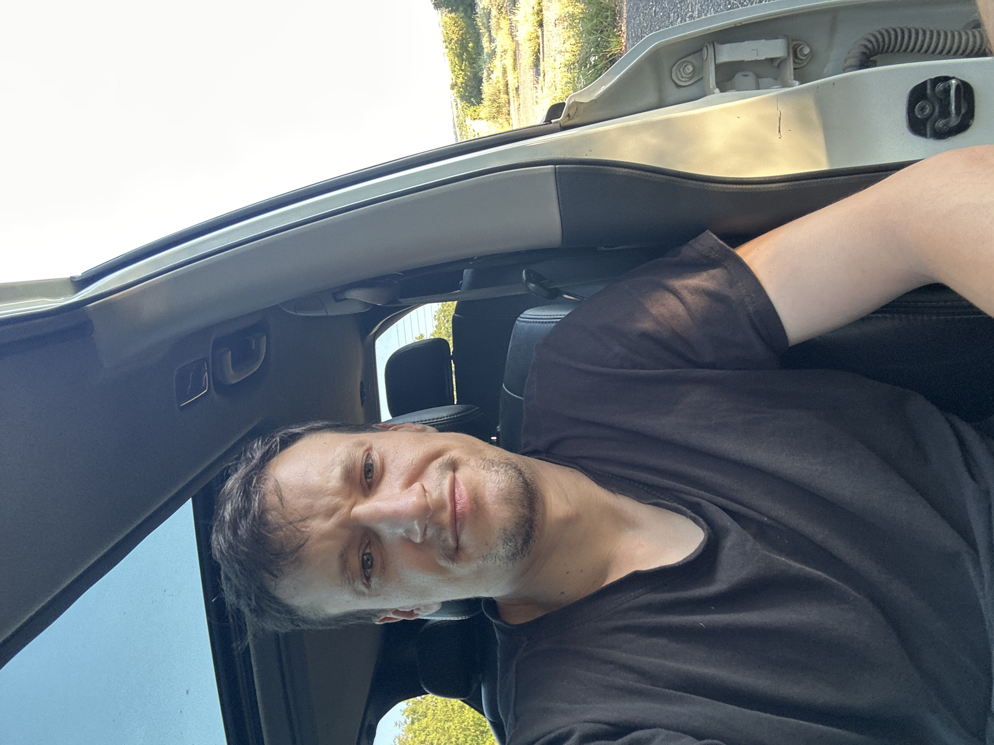 A man with short, dark hair and stubble is sitting in the driver's seat of a vehicle. He is wearing a black t-shirt. The vehicle's sunroof is open, revealing a blue sky. The car door is open, and the exterior shows a rural landscape with green trees and a field under a bright sky. The man is looking directly at the camera, with a slight smile. The scene appears to be outdoors, possibly on a sunny day.