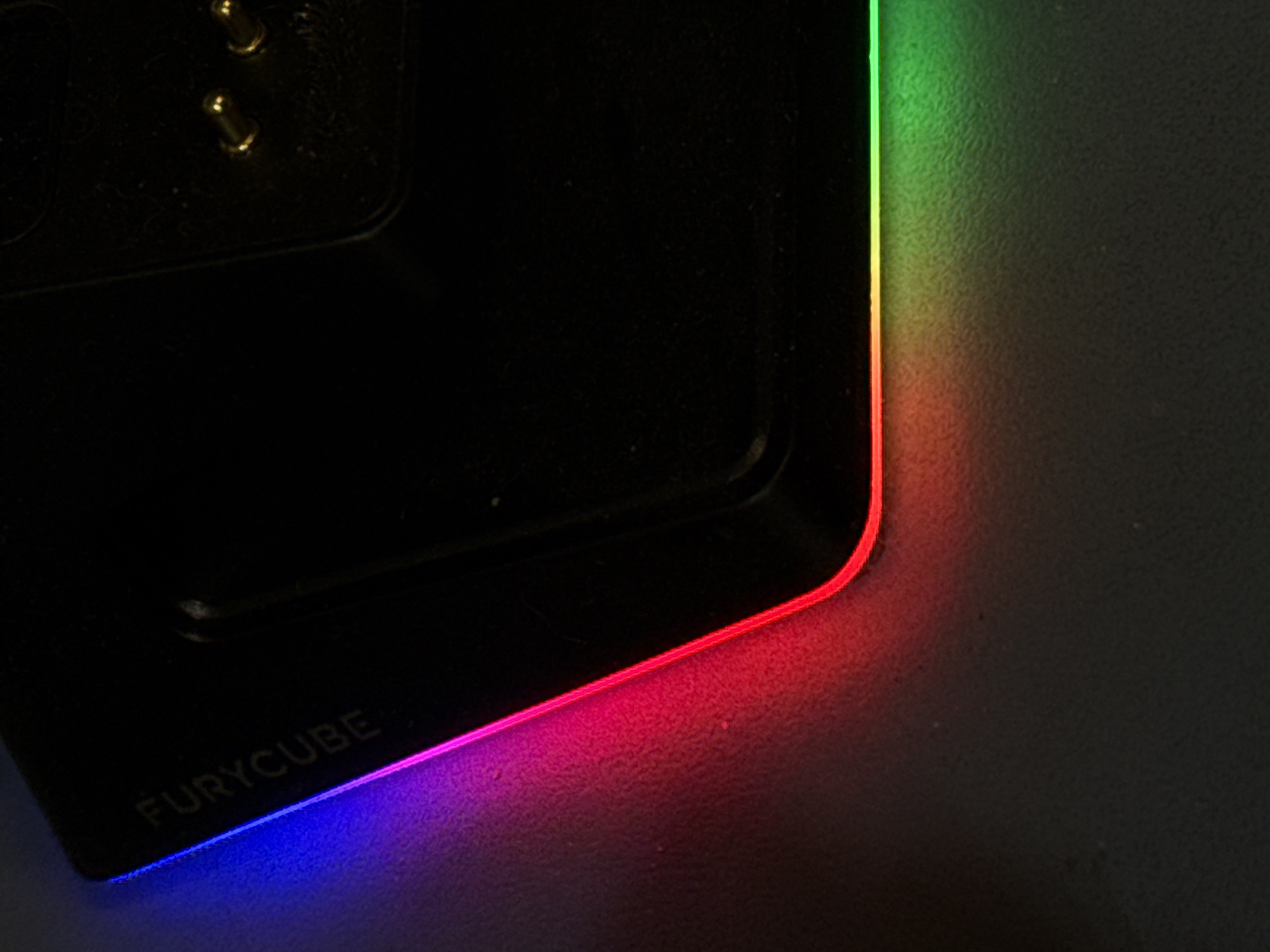 A close-up image shows a portion of a black Furycube device. The edges of the device have RGB LED lighting, transitioning from blue at the bottom left to red and then green along the top right edge. Part of the "FURYCUBE" logo is visible in the bottom left corner. The device rests on a textured gray surface. There is no location information in the image. No people, interactions, or actions are present.