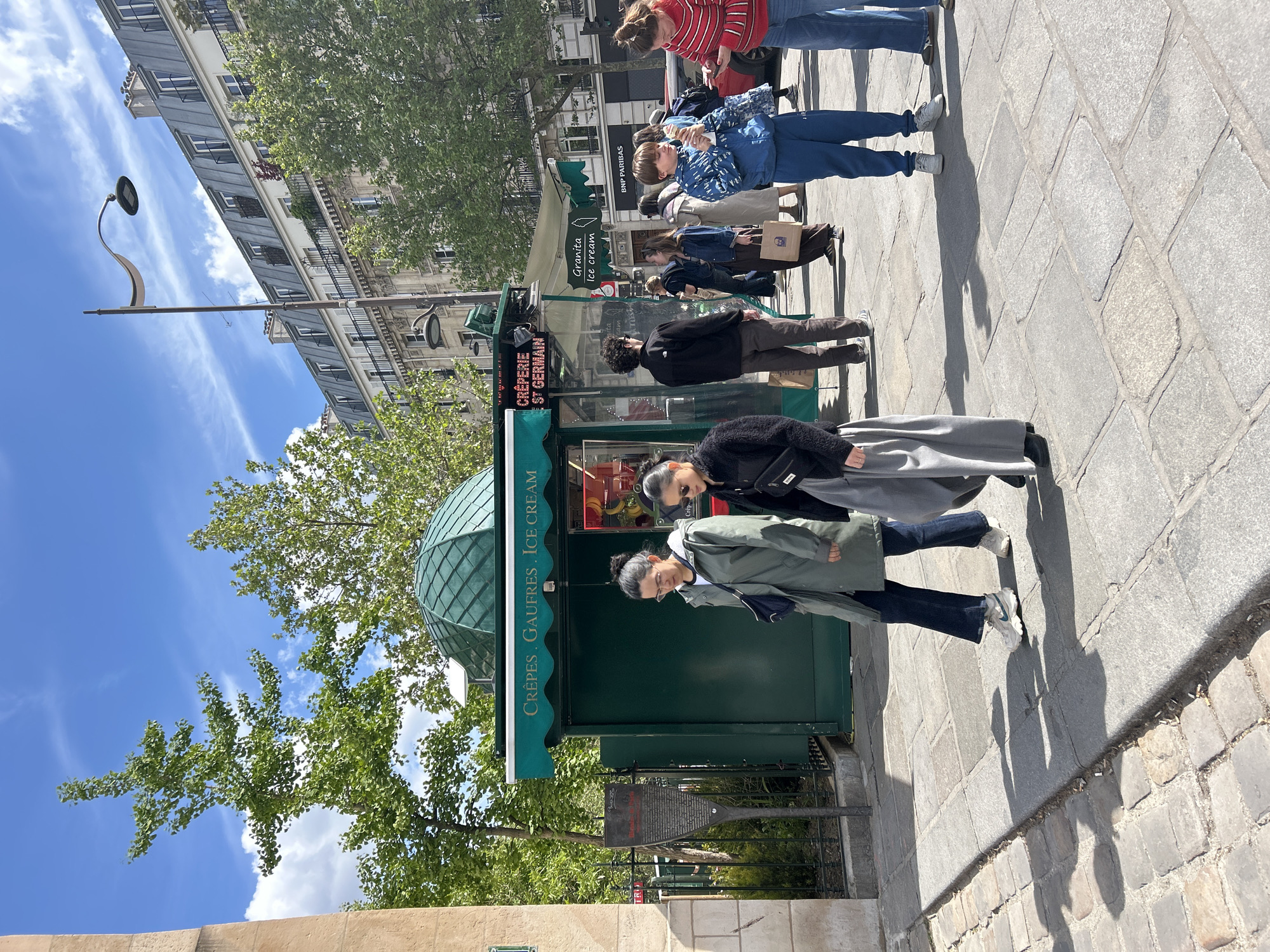 The image depicts a street scene with a Crêperie in St. Germain, Paris. People are seen standing near the stall, and a sign indicates that they sell Crêpes, Gaufres, Ice Cream, and Granita Ice Cream. A BNP Paribas bank is visible in the background. The environment includes trees, buildings, and paved streets typical of a Parisian urban setting.