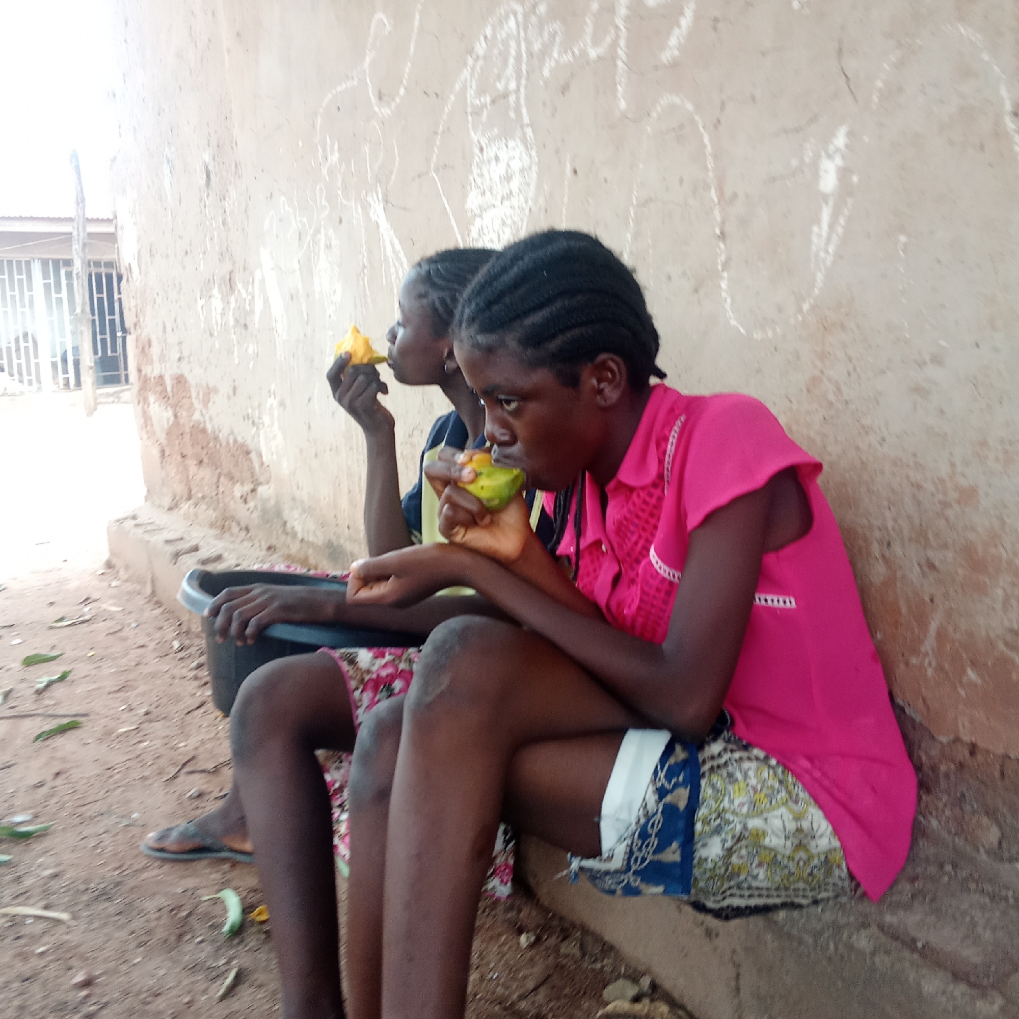 Two dark-skinned young women are sitting outdoors, eating fruits. The woman in the foreground wears a short-sleeved pink top and patterned shorts, while the other wears a blue top. A black container rests next to the first woman. They sit against a pale, weathered wall near a dirt ground with fallen leaves. A partially visible building with barred windows is in the background. The location is Akwanga, Nigeria.