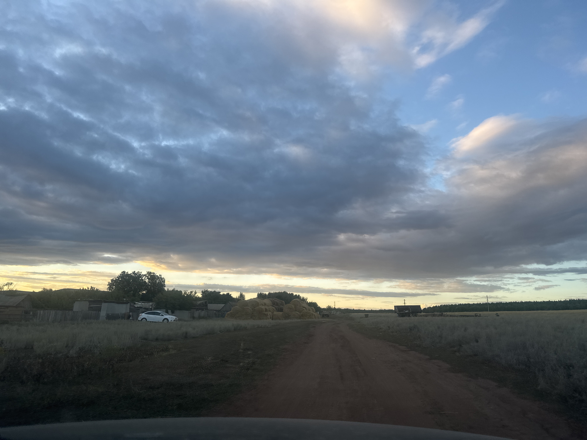 The image depicts a rural landscape in Vasilievka, Russia, under a dramatic sky with heavy, textured clouds ranging from dark gray to lighter, warmer tones near the horizon. A dirt road stretches into the distance, flanked by dry, sparse grass and vegetation. To the left, a cluster of weathered wooden structures forms a small settlement, with a white car parked in front of one of the buildings. Further into the scene, large piles of hay bales are visible in an open field, suggesting agricultural activity. The overall atmosphere is serene and slightly somber, with the setting sun casting a warm glow on the horizon, partially obscured by the clouds. The foreground shows the edge of a vehicle's interior, indicating the photo was taken from a car.
