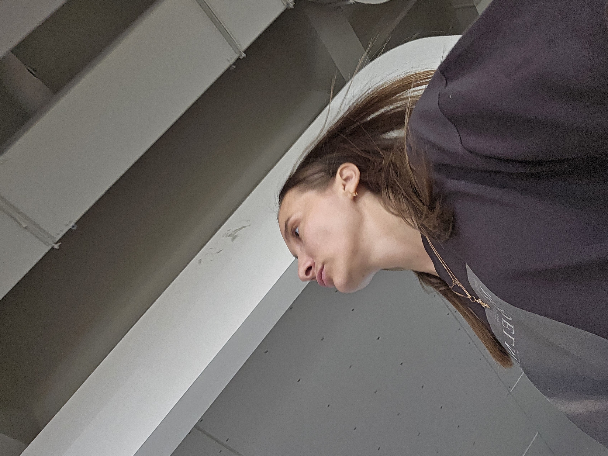 An upward-angled profile shot captures a young woman with her head tilted slightly forward and downwards, gazing to the viewer's left. Her long, medium-brown hair appears dynamically swept upwards and backwards, suggesting movement or a breeze. She is wearing a dark, possibly charcoal or navy, long-sleeved top, on which partial white text, appearing to read "JET", is visible on her right side. A delicate gold chain necklace with a small pendant hangs around her neck, and she has a small gold hoop earring in her visible ear. Her expression is neutral to slightly pensive, with her lips softly parted.
The background consists of stark, geometric structural elements, likely part of an indoor ceiling or modern architecture. These elements are wide, flat beams or ducts in shades of white and light grey, some exhibiting minor imperfections or dust. The lighting is even and diffused, indicative of an interior setting without direct sunlight. The overall composition places the woman prominently in the foreground, with the structured background providing a clean, minimalist context. The scene is situated in Chaikovsky, Russia.