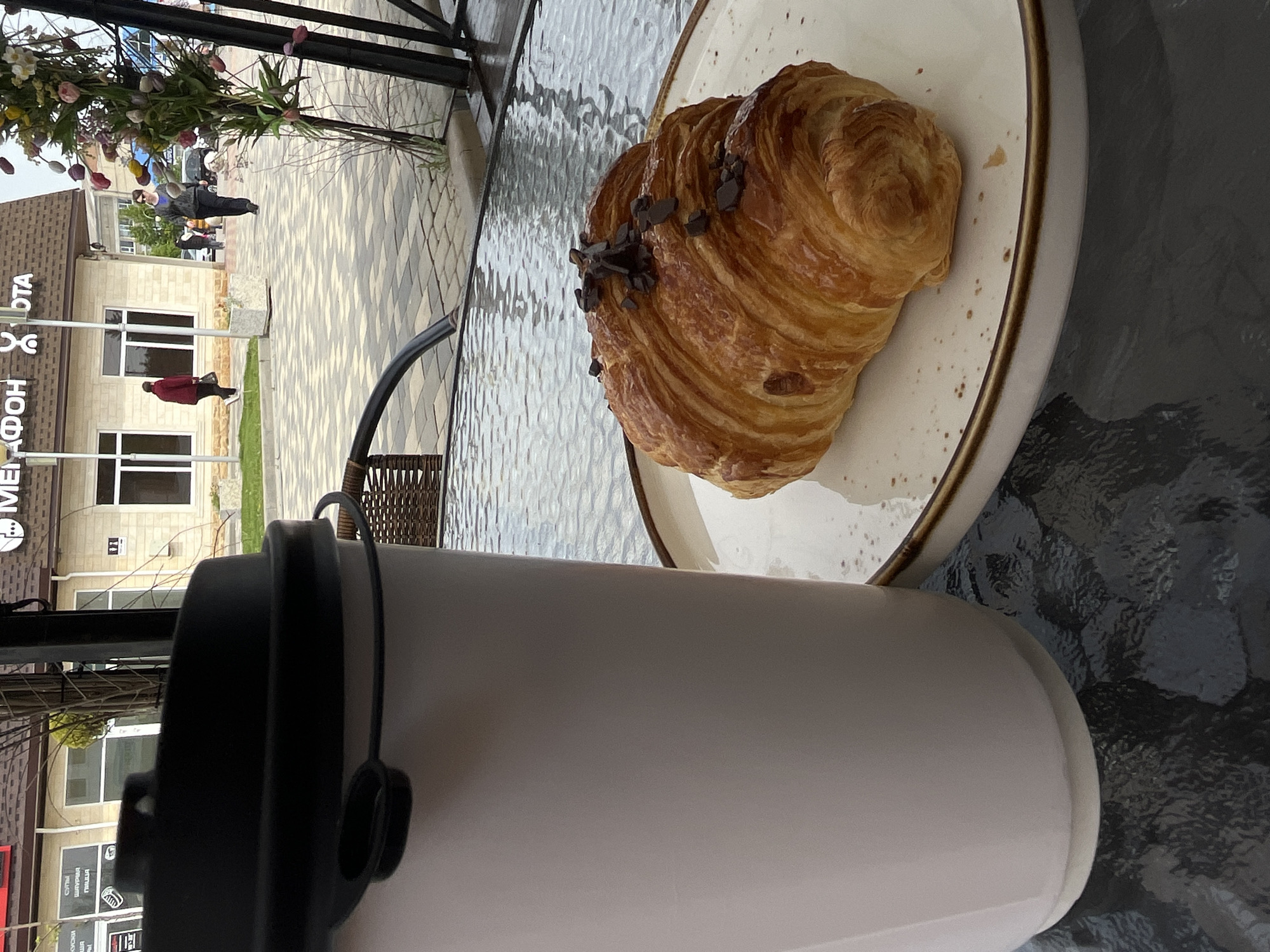 A high-angle shot shows a croissant on a plate next to a white cup on an outdoor table with a glass top. The croissant is topped with dark chocolate pieces. Beyond the table is a partial view of a building with Cyrillic script. People are walking on the sidewalk. The location is Novopokrovskaya, Russia. The focus is on the food and drink on the table.
