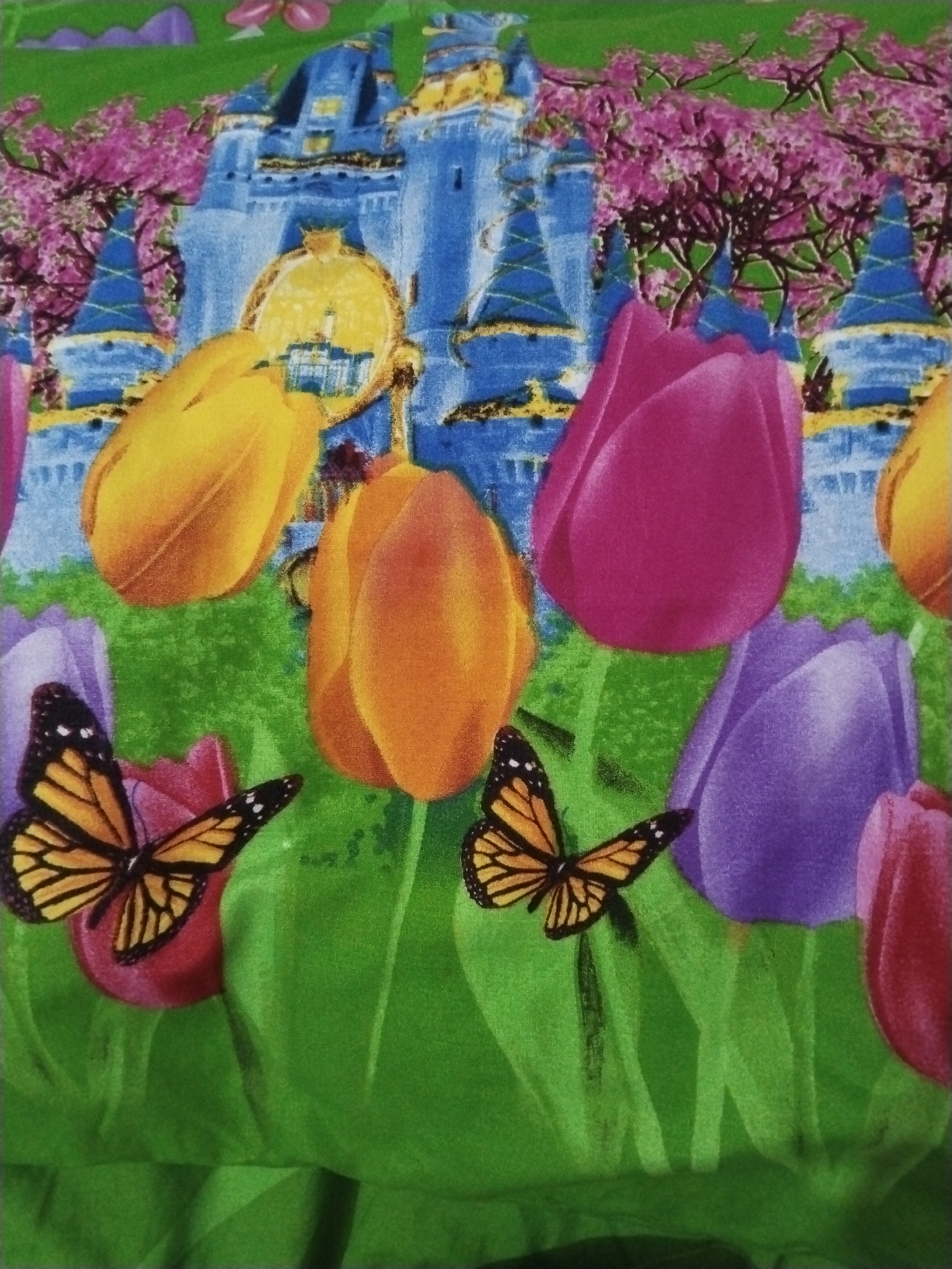 The image displays a colorful, illustrative scene featuring a blue castle with yellow accents, surrounded by vibrant pink trees and green foliage. In the foreground, several large, stylized tulips in shades of yellow, orange, pink, and purple are visible. Two monarch butterflies with black and orange wings are positioned on the tulips and foliage. The overall style is reminiscent of a children's book illustration or a fabric print. There is no discernible indication of location within Lawatan, Indonesia.