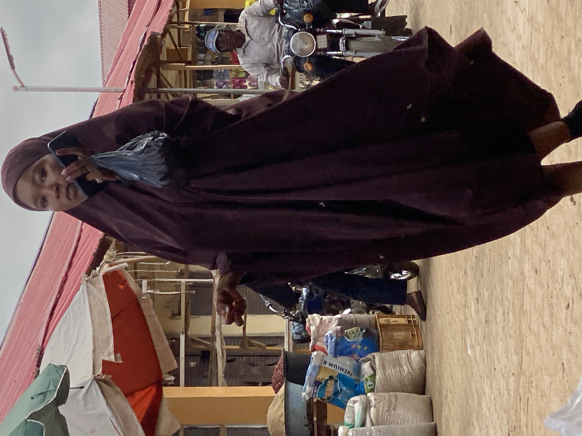 A woman in a hijab holds a smartphone in her left hand as she looks at the camera in Kafanchan B, Nigeria. She wears a long maroon dress. Behind her, a man wearing a blue cap and light blue shirt drives a motorcycle. Sacks of goods are stacked by a store. A red roof and an umbrella tent are visible.