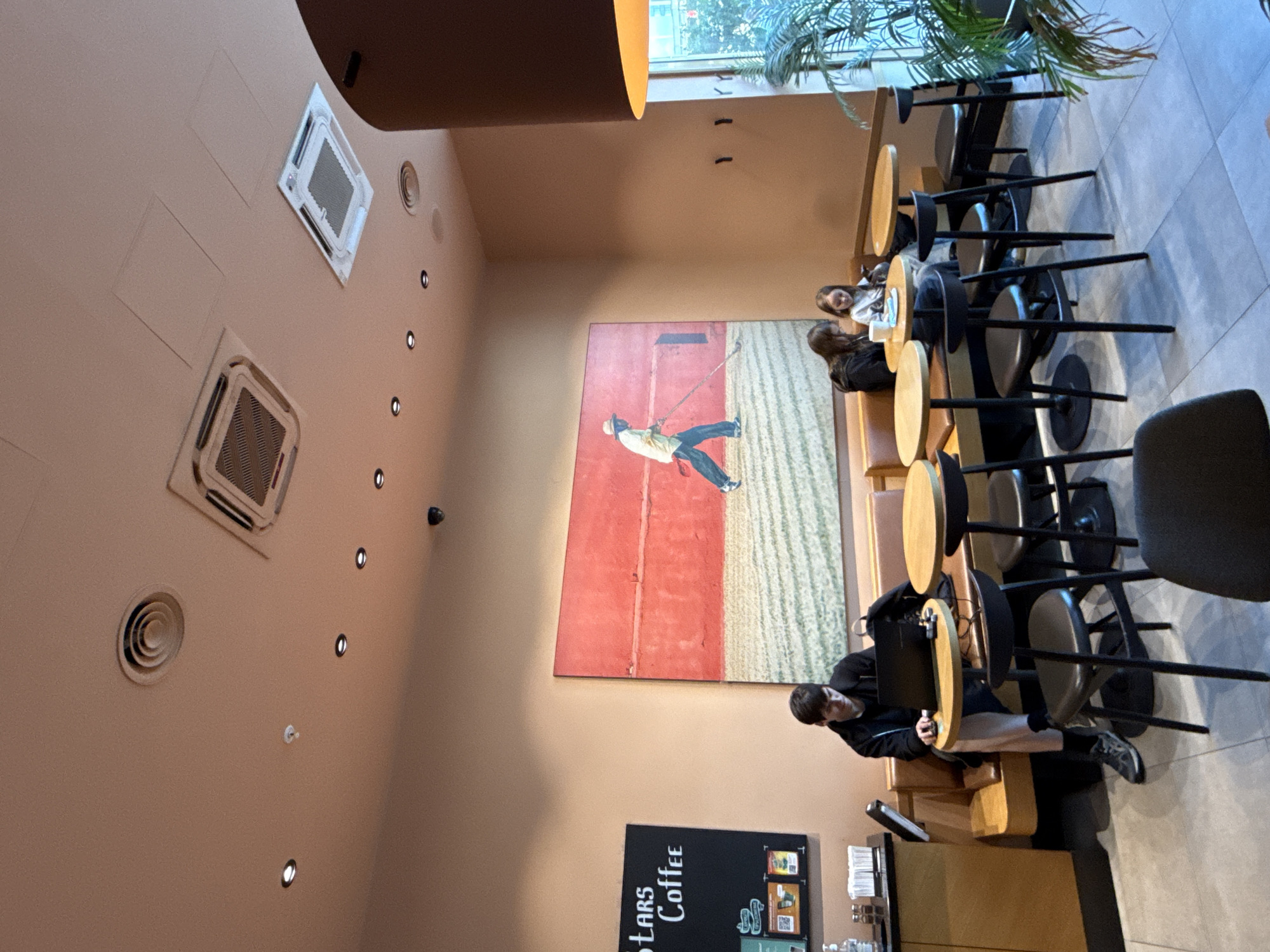 The image depicts the interior of a Starbucks coffee shop in Moscow, Russia. The scene is captured from a slightly elevated perspective, looking across the seating area. In the foreground, several small, round, light-wood tables with dark chairs are arranged. A large painting of a person in a hat raking a field against a bright red background hangs prominently on the wall.
To the left, a man is seated at a table with a laptop and a mouse, appearing to work or study. He is wearing a dark jacket and light-colored pants. A menu board with the text "STARS Coffee" is visible behind him. Further to the right, two young women are seated at another table, one looking at her phone and the other appearing to be engaged in conversation or also using her phone.
The environment includes a light-colored ceiling with recessed lighting and air conditioning vents. A large, dark brown lampshade hangs from the ceiling on the right side of the frame, and a potted plant with green fronds is situated near the window on the far right. The overall ambiance suggests a typical coffee shop setting with customers engaged in various activities.