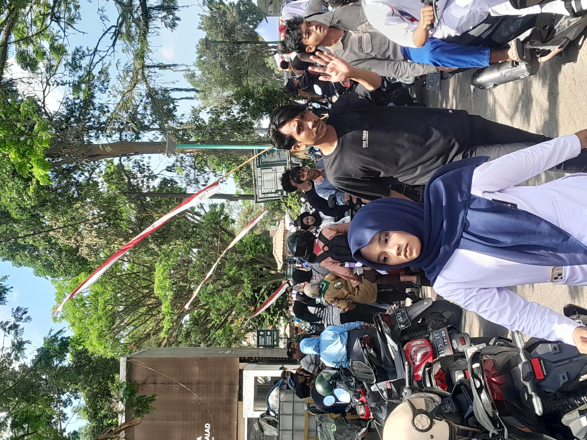 The image depicts a street scene in Genteng, Indonesia, possibly near a parking area or a community gathering. In the foreground, a young woman wearing a blue hijab and a white long-sleeved shirt is looking to her left. To her right, a man in a black t-shirt with the text "USA TEAM CHAMPIONSHIP" is waving with his right hand and looking towards the camera. Behind them, several other people, motorcycles, and trees are visible. Indonesian flags are strung across the street, suggesting a celebratory or patriotic event. Signs indicating parking for cars and motorcycles, as well as a toilet, are present on a pole in the background. The overall atmosphere appears to be a bustling outdoor environment on a sunny day.