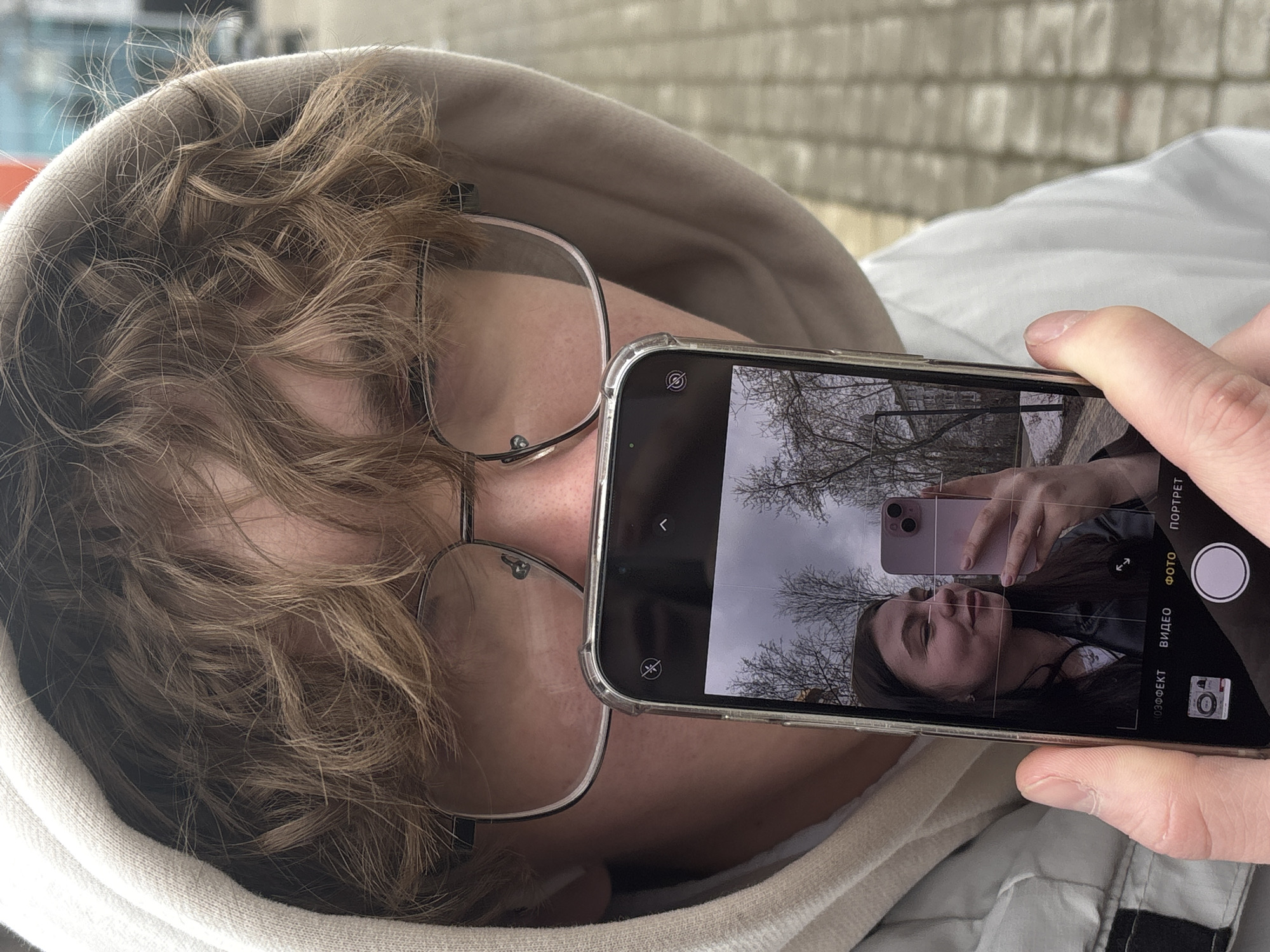 A person with light brown curly hair and glasses, wearing a beige hoodie, is holding a smartphone. The smartphone screen shows the reflection of a woman taking a selfie with a pink smartphone. The background in the smartphone reflection includes bare tree branches and an overcast sky. The person holding the phone has their face partially visible behind the phone. The smartphone interface is set to camera mode, with Russian text visible on the side. The location is suggested to be in Perm, Russia. A brick wall is in the background.