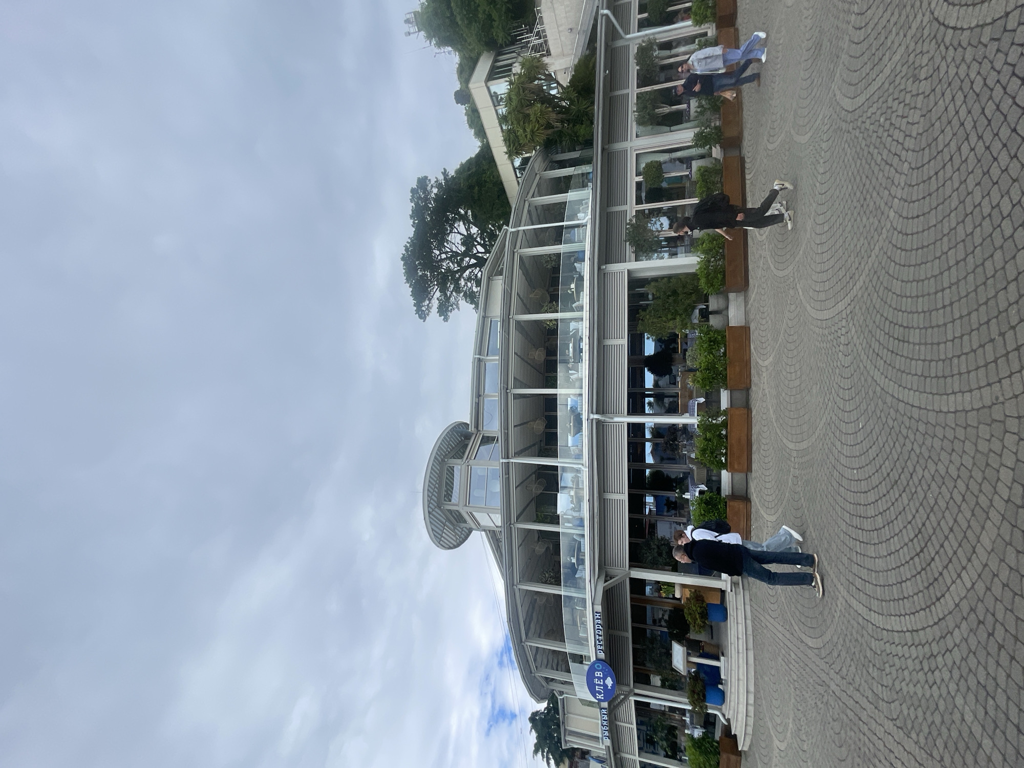 The image shows a two-story, modern glass-enclosed restaurant building with a circular design on a paved area. Several people are walking on the sidewalk. A sign with the words "рыбный КЛЕВО ресторан" (fish restaurant CLEVER) is visible in Russian. The sky is overcast. The location is in the city of Sochi, Russia.