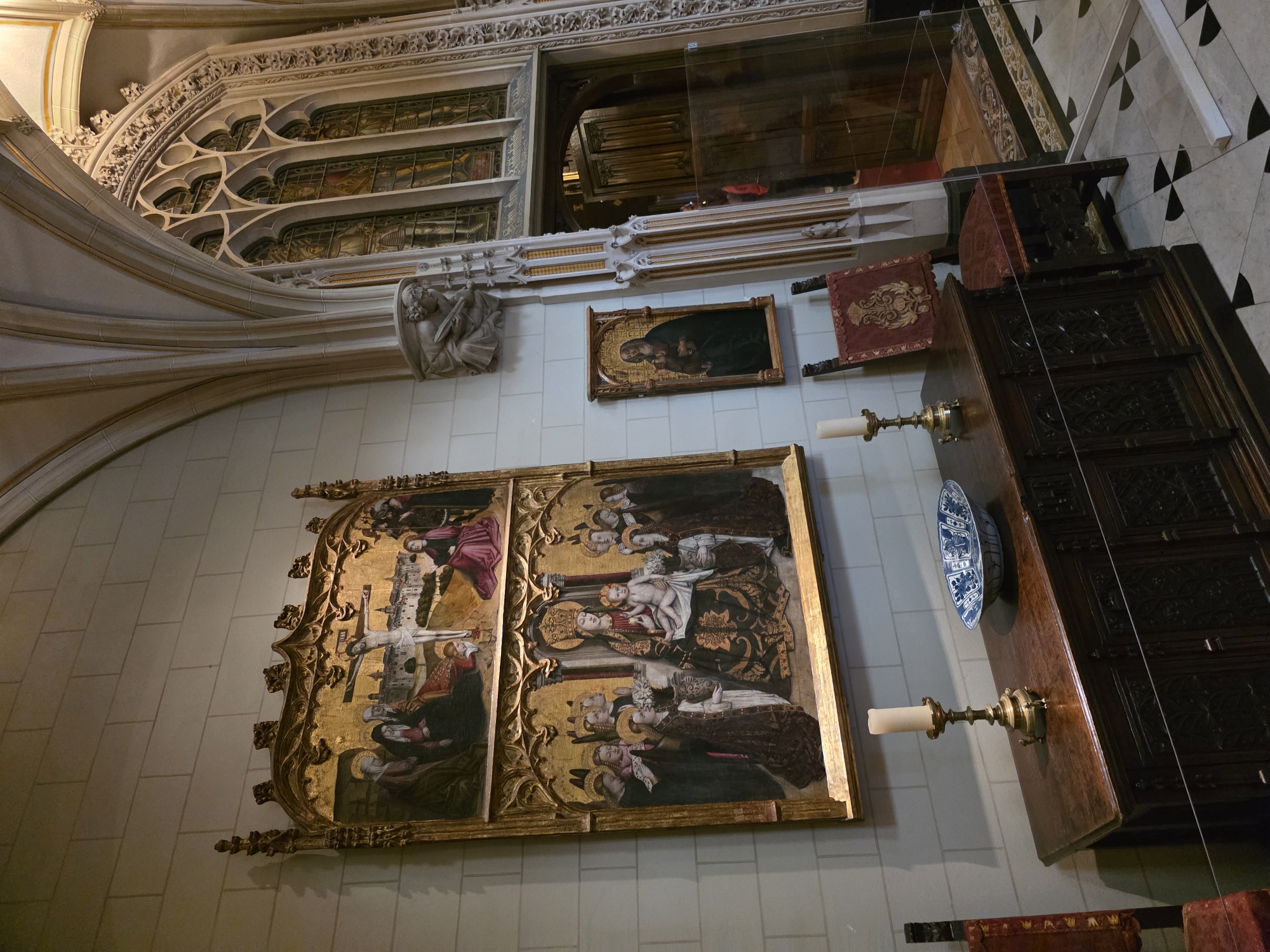 The photograph captures an interior view of a Gothic-style room, likely within a historical building. Dominating the scene are two large, ornate religious paintings displayed on a pale, textured wall. The upper painting depicts the crucifixion of Jesus, with figures of Mary and John present. The lower painting shows the Virgin Mary enthroned with the infant Jesus, surrounded by saints. These paintings are framed in elaborate gold leaf.
In the foreground, a dark, intricately carved wooden chest of drawers serves as a display surface. On top of the chest are two brass candlesticks with lit candles and a blue and white ceramic bowl. To the left of the chest, part of a red upholstered chair is visible. To the right of the chest, a separate, smaller framed painting of the Madonna and Child hangs on the wall.
The architecture features pointed arches, ribbed vaulting, and decorative stonework. To the right, a grand arched doorway leads into another room, partially obscured by a glass barrier. Stained glass windows, depicting figures in armor, are visible through the arched opening of the doorway. The floor is a black and white checkered pattern.
The scene is static, with no visible individuals actively engaged in any interactions within the frame, although figures are depicted within the paintings. The lighting appears to be a mix of natural light filtering through windows and artificial light from the candles and possibly unseen sources, casting shadows and highlighting the details of the artwork and furnishings.
Based on the architectural style and the presence of religious art, this location is likely a museum or a historically significant religious building. However, without further context or external information, it is not possible to definitively place this scene within a specific location in Utrecht, Netherlands. The image solely depicts the interior of this room.