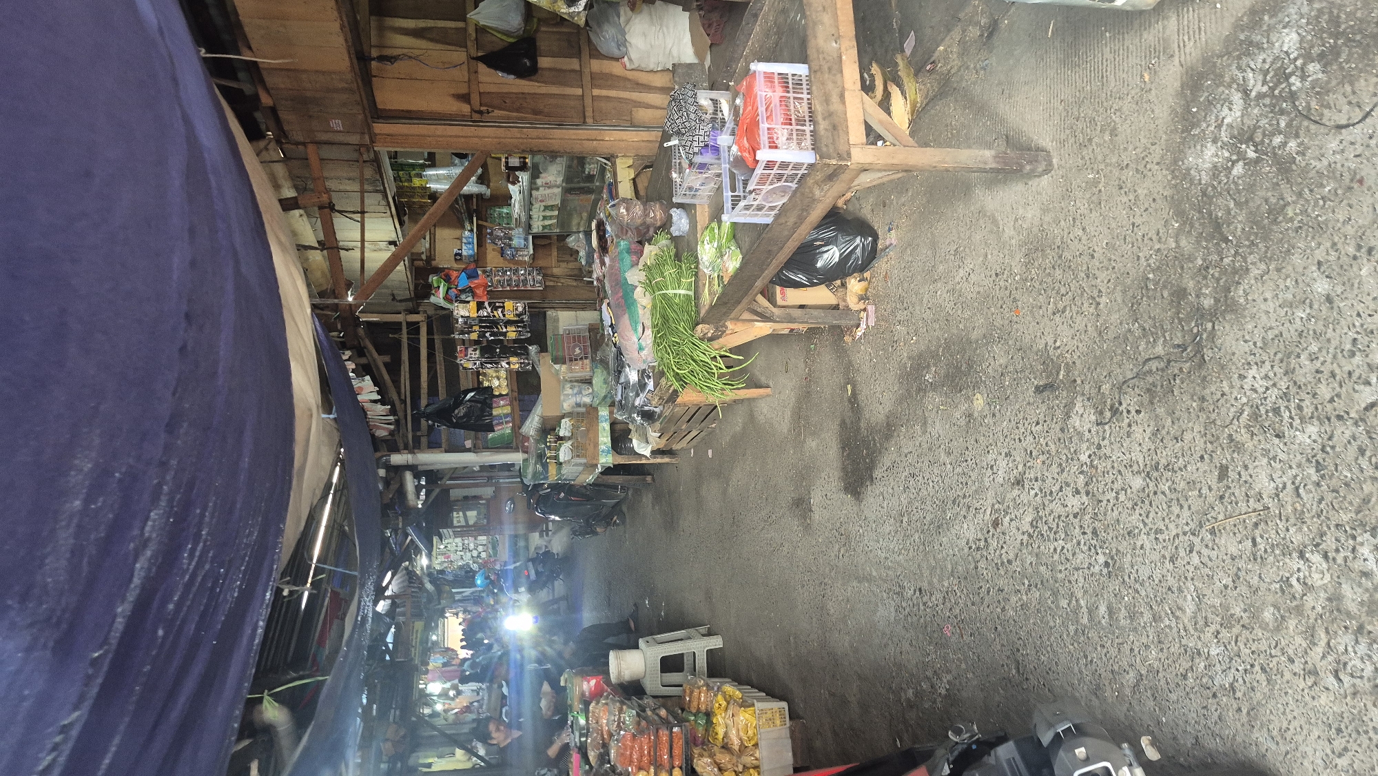 The image depicts a street-level view of a market in Boja, Indonesia. A large blue tarp covers part of the scene, providing shade. The market is lined with stalls made of wood and crates, displaying various goods. In the foreground, a wooden table holds a bundle of long green beans and some leafy green vegetables. To the right, on another wooden table, are two white plastic crates, one containing red items and the other with a patterned fabric. In the background, people are visible, some tending to stalls and others walking through the market. Motorcycles are parked in the aisle, and goods are stacked on shelves and tables. The ground is paved with concrete. The overall impression is of a busy, open-air marketplace.