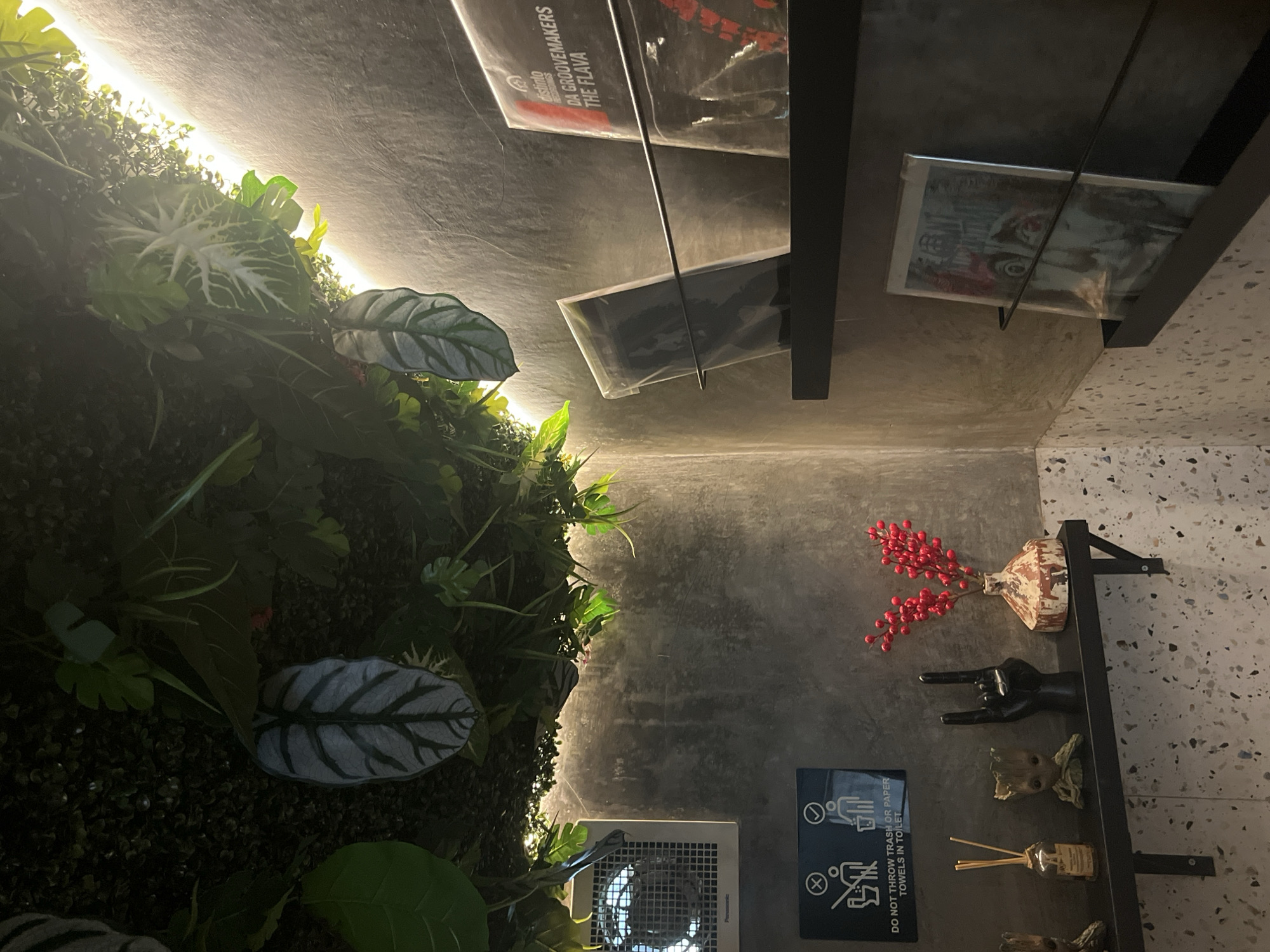 The image depicts a modern, dimly lit interior space, possibly a restroom or a niche area within a commercial establishment in Kuta Selatan, Indonesia. Dominating the upper portion of the frame is a lush, artificial plant installation, featuring a variety of green leaves and foliage, accented by warm, ambient lighting that casts a glow from the right side. The walls are textured concrete, with a lighter, speckled material lining the lower portion of the wall and continuing onto a shelf.
On the left wall, a white ventilation fan is mounted. Below it, a black sign with white icons and text reads, "DO NOT THROW TRASH OR PAPER TOWELS IN TOILET," indicating the function of the space.
A black shelf is mounted on the wall. On this shelf, from left to right, are several decorative items: a small bottle with diffusers, a miniature Groot figurine, a black hand gesture sculpture making the "rock on" sign, and a small, distressed-look vase filled with bright red berries.
Above this shelf, on another black shelf, are several vinyl records displayed upright in sleeves. The topmost record on the right is clearly visible with text that reads "DISTINLO Records" and "DA GROOVEMAKERS THE FLAVA." Another record is partially visible to its left, and a third record is displayed on a shelf below, featuring a face on its cover. The overall impression is one of a stylishly decorated, contemporary space with a focus on ambiance and curated aesthetics.