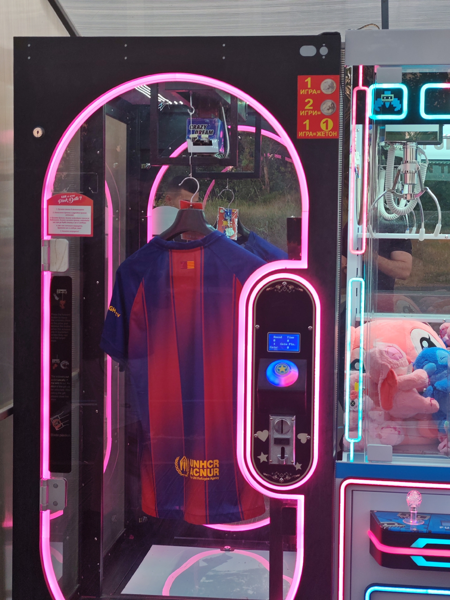 This photograph, taken in Nesebar, Bulgaria, shows a claw machine with a Barcelona soccer jersey as a prize, which is hung on a hanger inside the claw machine. The machine features pink and blue neon lights and a coin slot. A sign indicates the cost of play, with one game costing two coins or one token, while another sign provides instructions. Stuffed animals are visible in the right section of the machine.