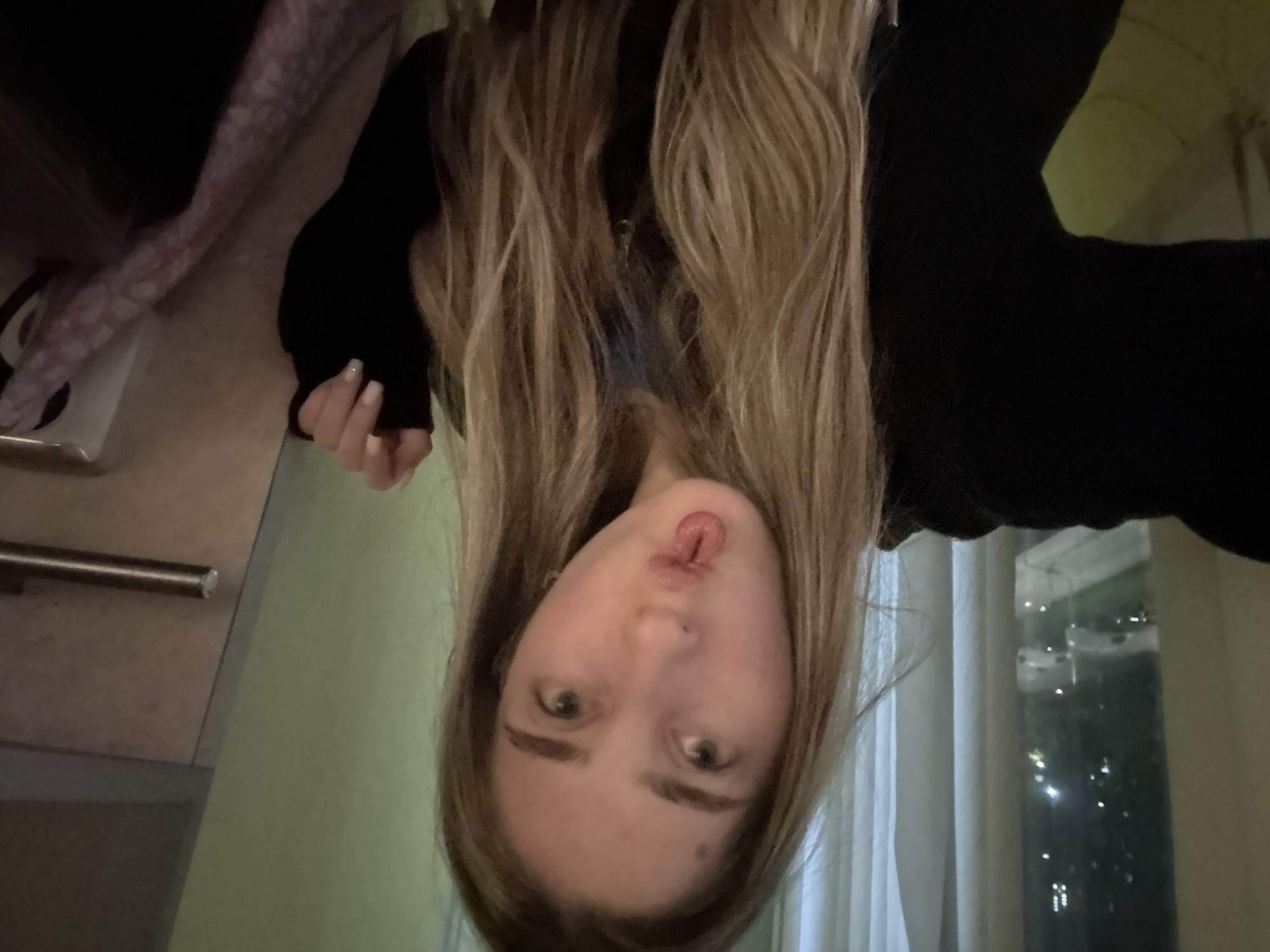 A person with long, blonde hair and wearing a black long-sleeved shirt is upside down in a room. They have fair skin, light-colored eyes, and are sticking out their tongue slightly. In the background, there is a window with dark visibility outside and some visible city lights. To the left, there is a cabinet with a metal handle and a light green wall. There is also a pink and purple patterned fabric. The location is an unspecified city.
