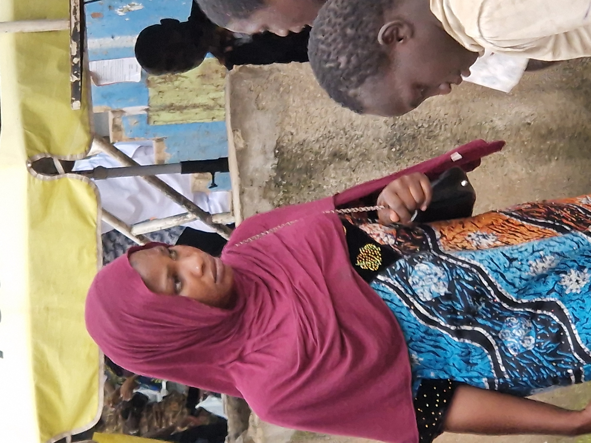 A medium shot captures a young Nigerian woman wearing a dark red hijab and a colorful, patterned dress. She is looking to her right, with a serious expression. To her right, a young boy in a light-colored hoodie is visible from the side, looking down. Another individual, whose face is obscured by shadow, is standing further in the background to the right. The background includes a yellow awning, a blue and light green painted wall, and a concrete structure. The scene appears to be outdoors, likely in a market or street environment within the city of Jos, Nigeria. The lighting is somewhat dim, suggesting it could be overcast or late in the day. The overall impression is one of everyday life in a bustling urban setting.