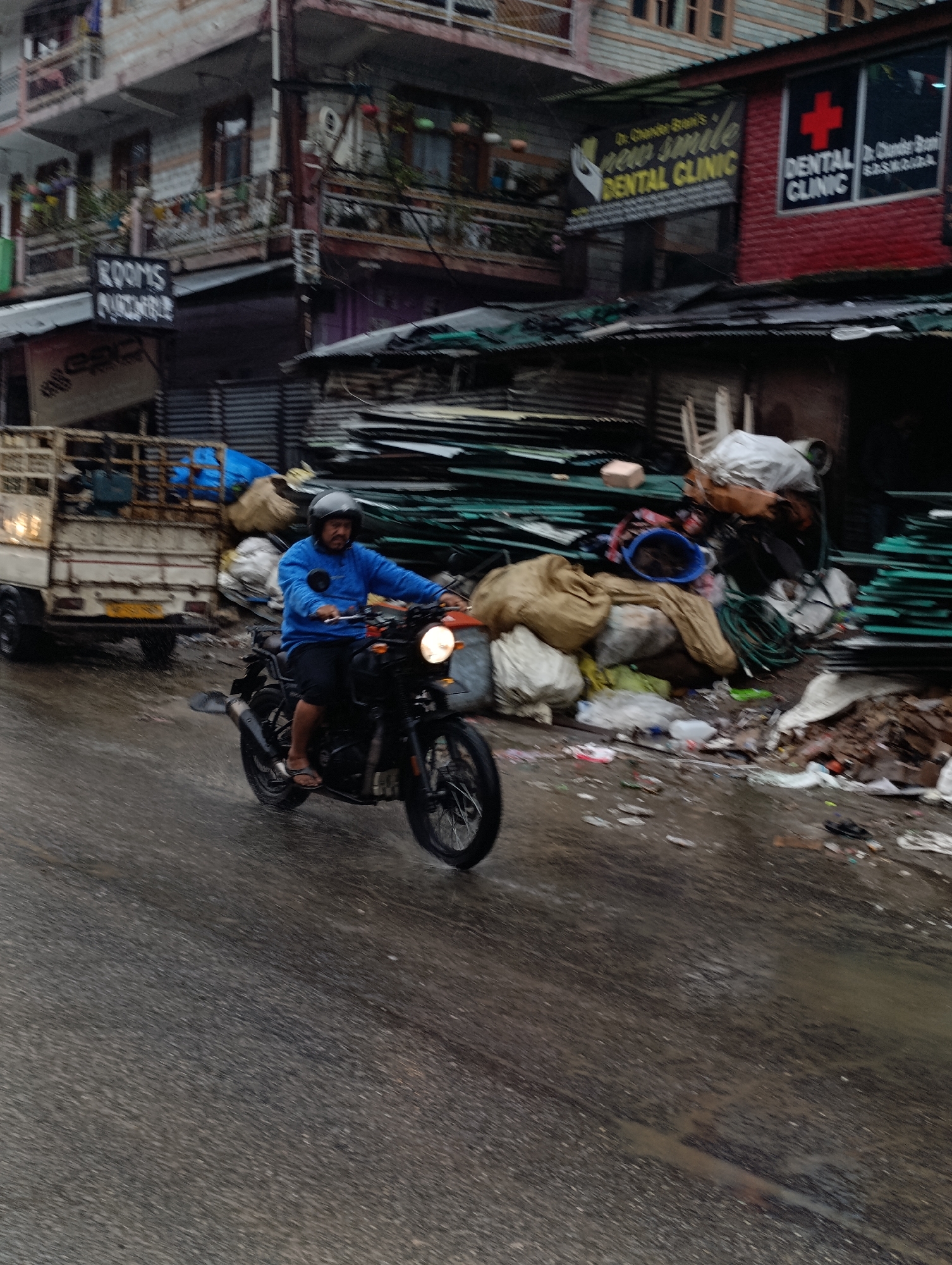 A man wearing a blue shirt, black shorts, and a black helmet rides a black motorcycle on a wet, paved street in Manali, India. The motorcycle has a round headlight and black wheels. In the background, there is a multi-story building with balconies. To the right of the building, a red sign with a white cross and text that reads "DENTAL CLINIC" is visible. Adjacent to it, a banner with yellow text proclaims "new smile DENTAL CLINIC". Further in the background, a white truck is parked on the left side of the street. Piles of assorted materials, including sacks, plastic sheeting, and what appears to be debris, are stacked along the roadside. The road surface is wet, indicating recent rain.