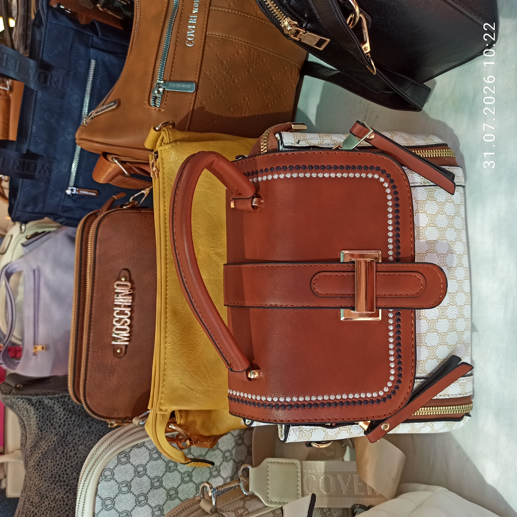 The image shows a display of handbags in a retail setting. The handbags are of various colors and styles, including brown, yellow, navy blue, and black. Some of the handbags have brand names such as "MOSCHINO" and "COVERI WORLD" visible on them. The scene appears to be indoors, likely a shop, with a white surface in the foreground. The image also includes a timestamp in the bottom right corner: "31.07.2026 10:22". There are no people visible in the image. The location in Modugno, Italy is not discernable from the image itself.