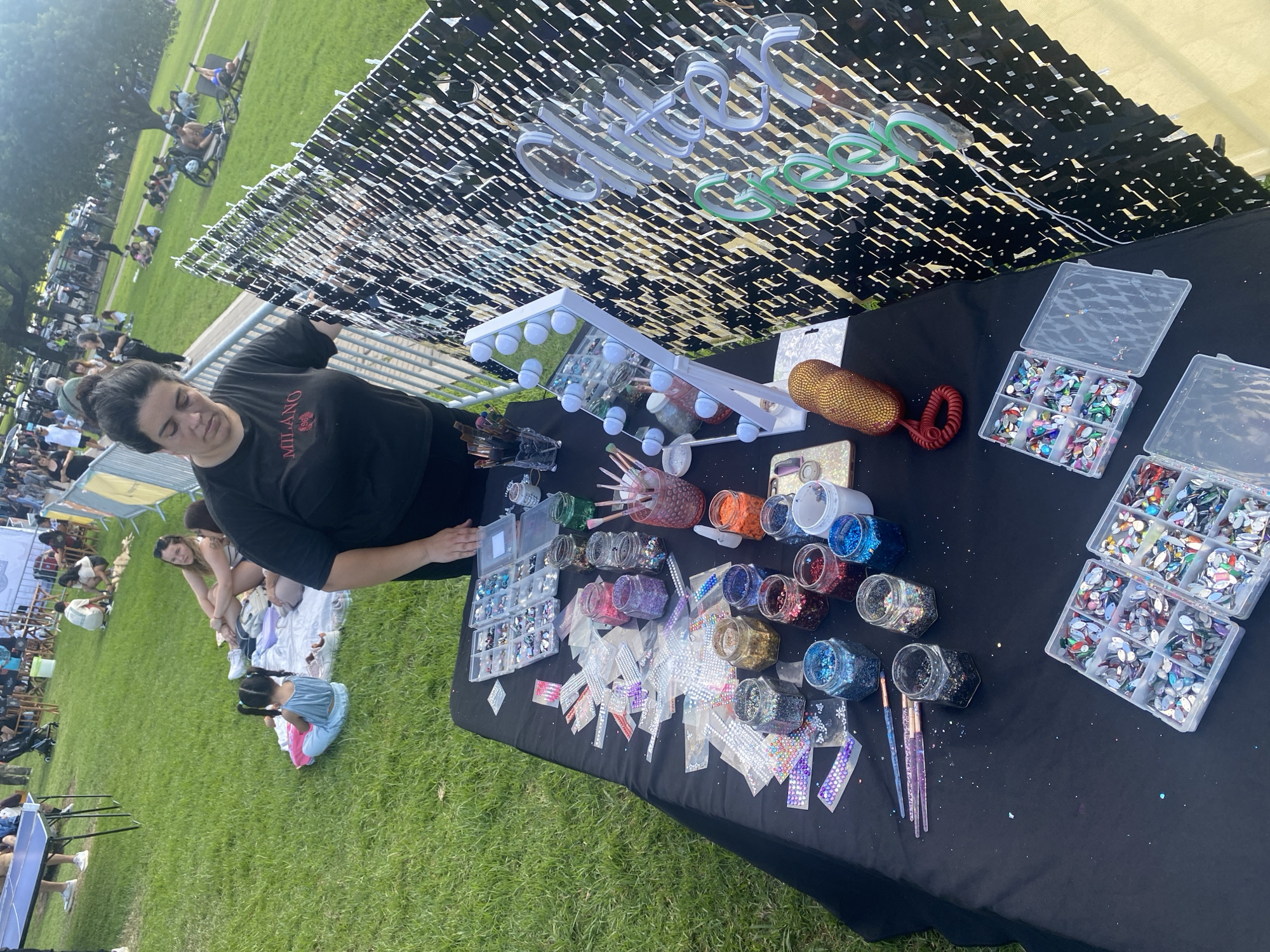 The image shows a woman standing behind a table covered in glitter, rhinestones, and various other craft supplies. A sign that reads "Glitter Green" hangs above the table. The woman is wearing a black shirt with the word "Milano" on it. In the background, there is a large open grassy area with people sitting and relaxing. This is most likely in the city of Buenos Aires, Argentina, though specific landmarks or details to confirm this are not visible. The woman is likely working at a craft fair or market.