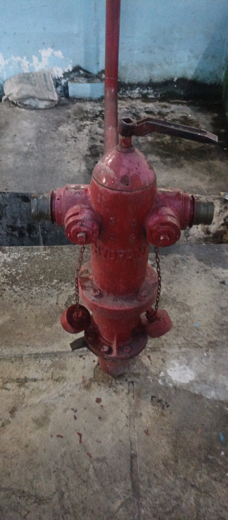 The image depicts a red fire hydrant situated on a concrete surface. The hydrant is upright and appears to be made of metal, with a central vertical pipe and a wider base. It has multiple outlets, each covered with a cap suspended by a chain. A handle mechanism is visible at the top, suggesting it is a functional water source. The surrounding environment appears to be an outdoor urban setting, possibly an alleyway or service area, with a textured concrete ground and a wall in the background painted in light blue with some peeling. Debris and dirt are scattered on the ground. The scene is illuminated by artificial light, casting shadows and highlighting the textures of the surfaces. The overall condition of the hydrant and its surroundings suggests it is in a functional but somewhat weathered state, consistent with typical urban infrastructure. There are no people visible in the image.