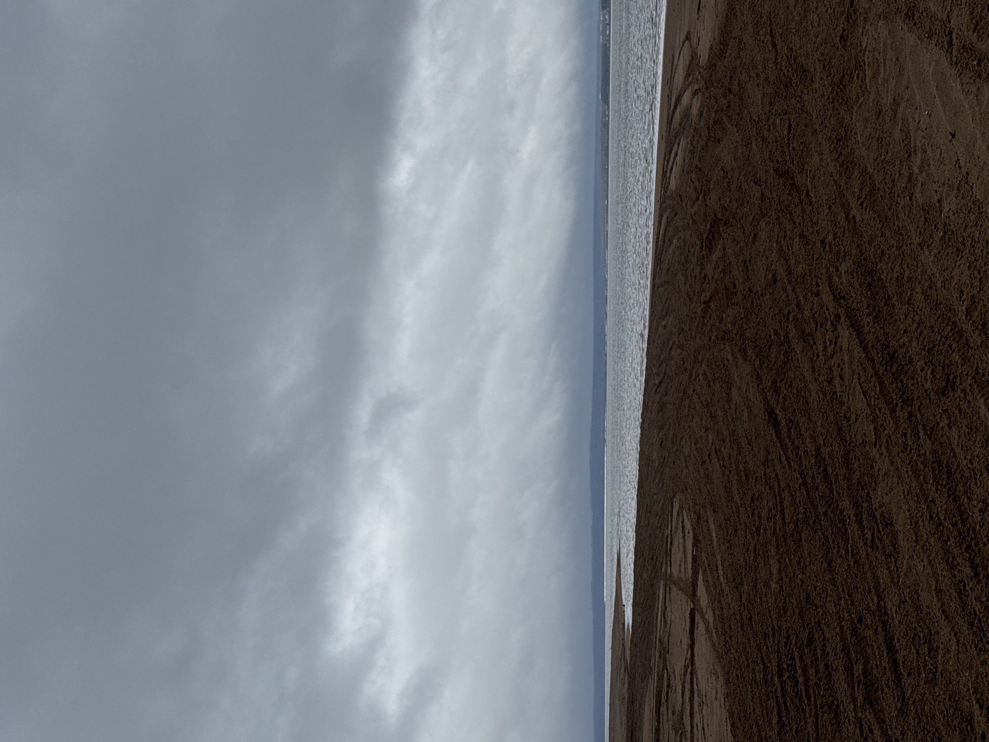 The image shows a beach scene in Noviy, Russia. The sky is overcast with gray and white clouds filling the left side of the frame. The sea is a strip of gray-blue with white foamy waves, and a faint outline of a town is visible in the distance. The right side of the frame is covered with dark brown sand. There are traces on the sand, indicating activity, but no people are visible in the frame. The image is taken from a low angle.